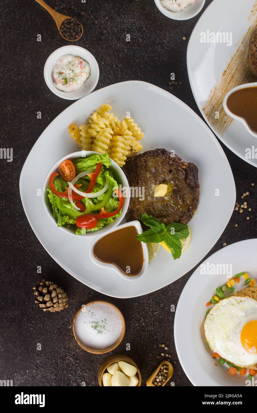 Bistecche di manzo alla griglia con patate, verdure e salsa di bistecca a piatto bianco su un tavolo rustico texture Foto Stock