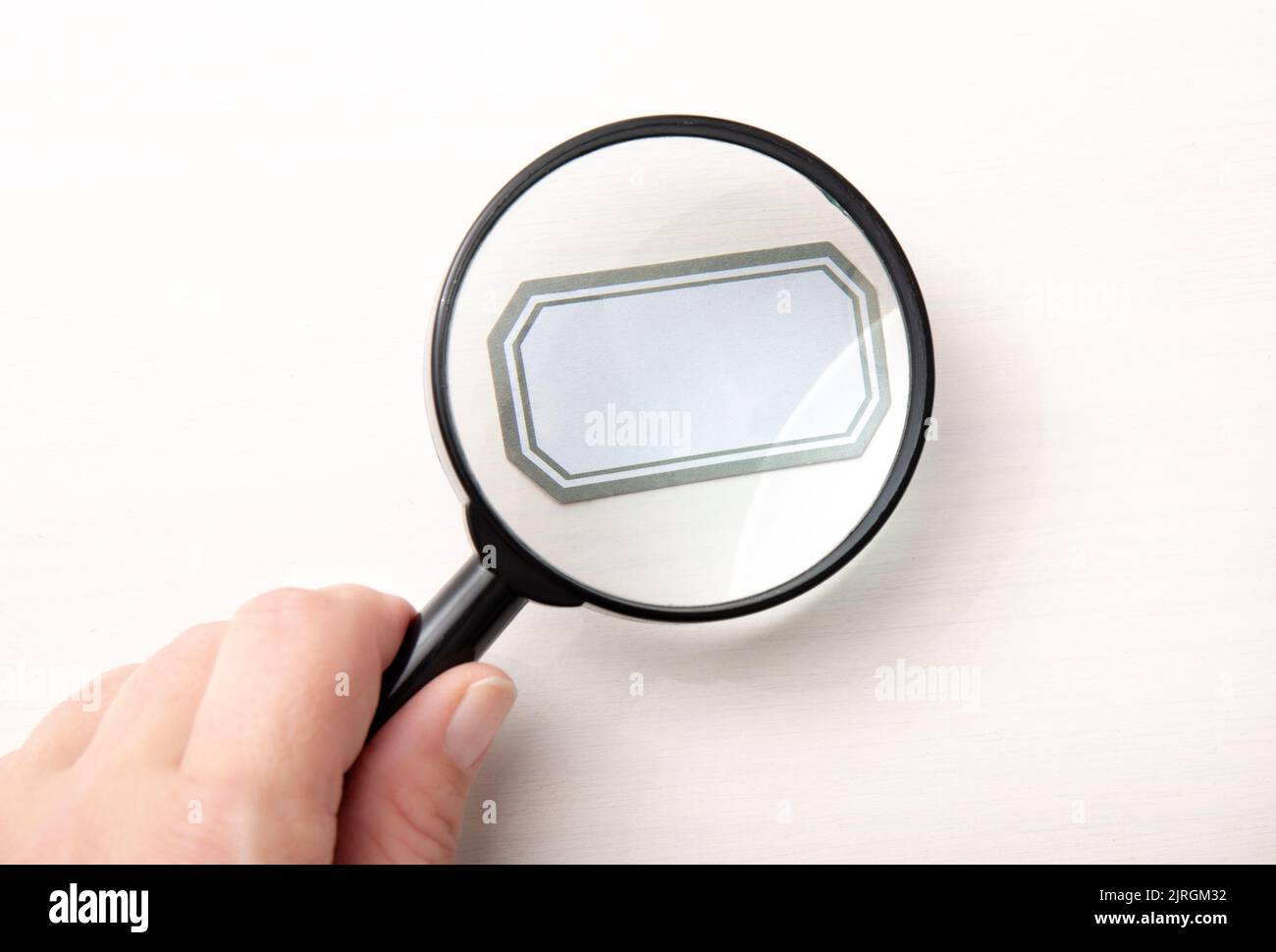 Mano della persona che tiene la lente d'ingrandimento tradizionale nera e che osserva l'etichetta vuota, sfondo bianco. Compilate voi stessi. Foto Stock