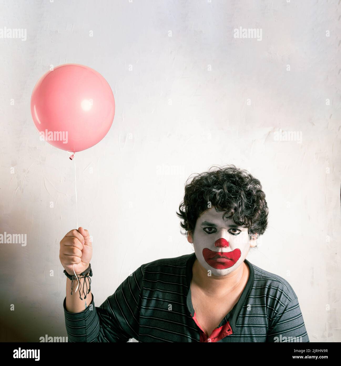 Un maschio riccio con una faccia di clown triste che tiene un palloncino rosa Foto Stock