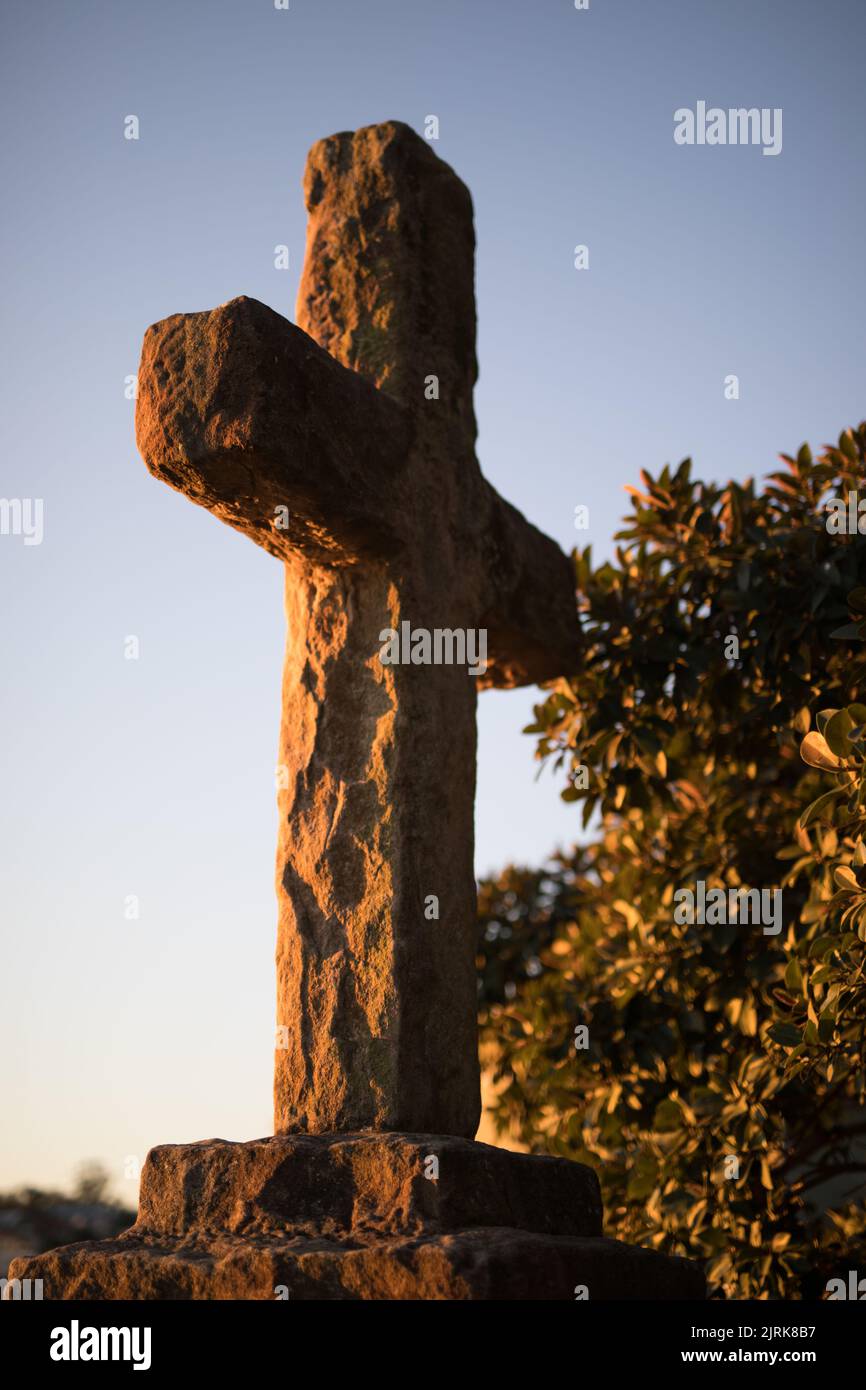 Un colpo verticale di una croce di pietra su un piedistallo contro alberi verdi all'ora d'oro Foto Stock