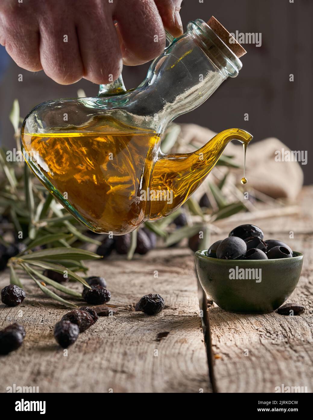 Chef irriconoscibile che versa olio fresco su olive nere mature sopra un tavolo di legno vicino ai rami Foto Stock