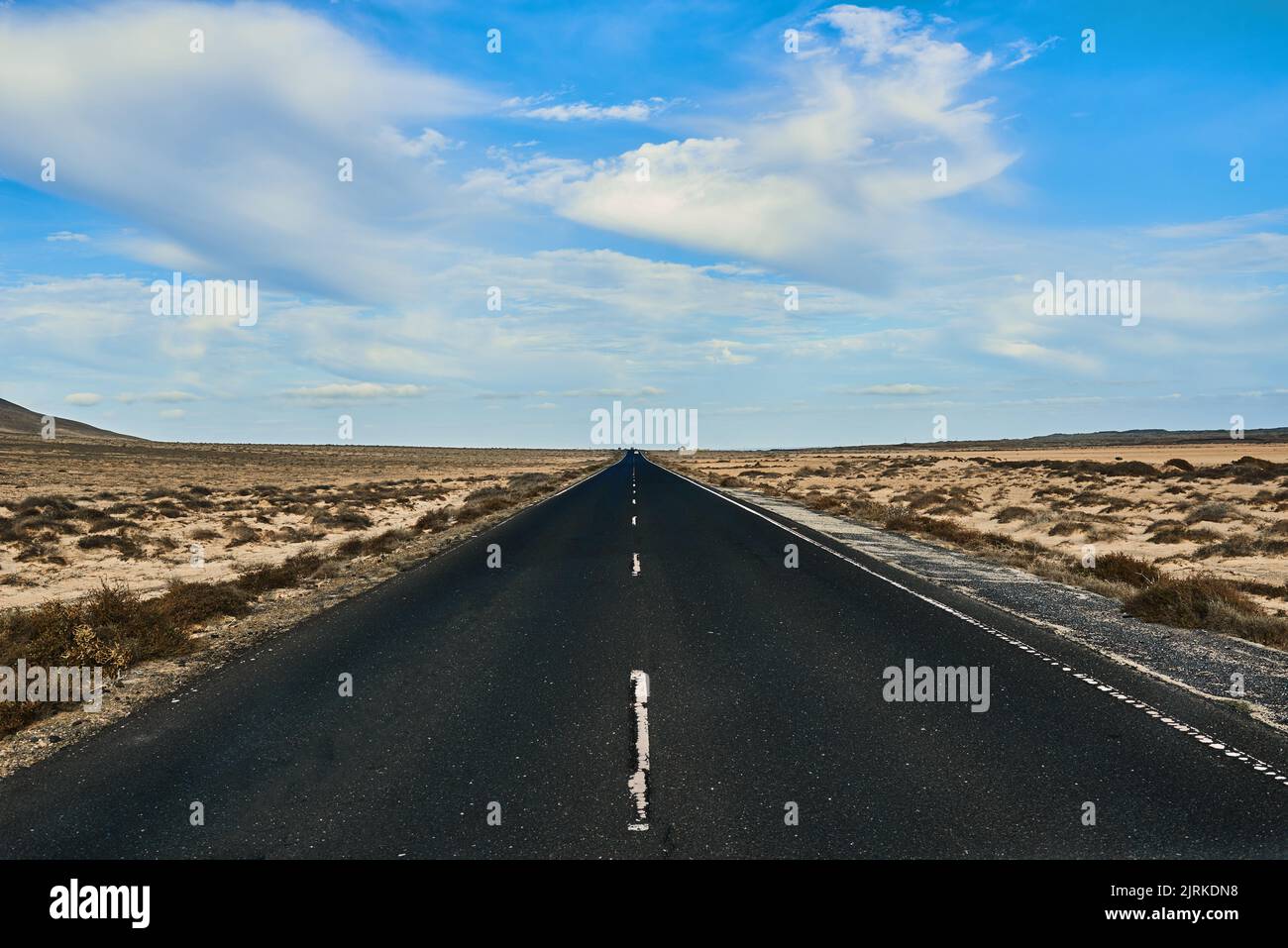 Strada diritta tra terra sabbiosa con piante e montagna sotto cielo nuvoloso blu a Fuerteventura Spagna Foto Stock