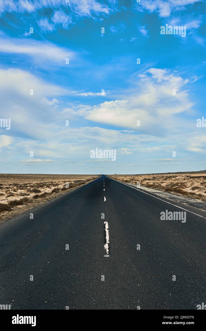 Strada diritta tra terra sabbiosa con piante e montagna sotto cielo nuvoloso blu a Fuerteventura Spagna Foto Stock