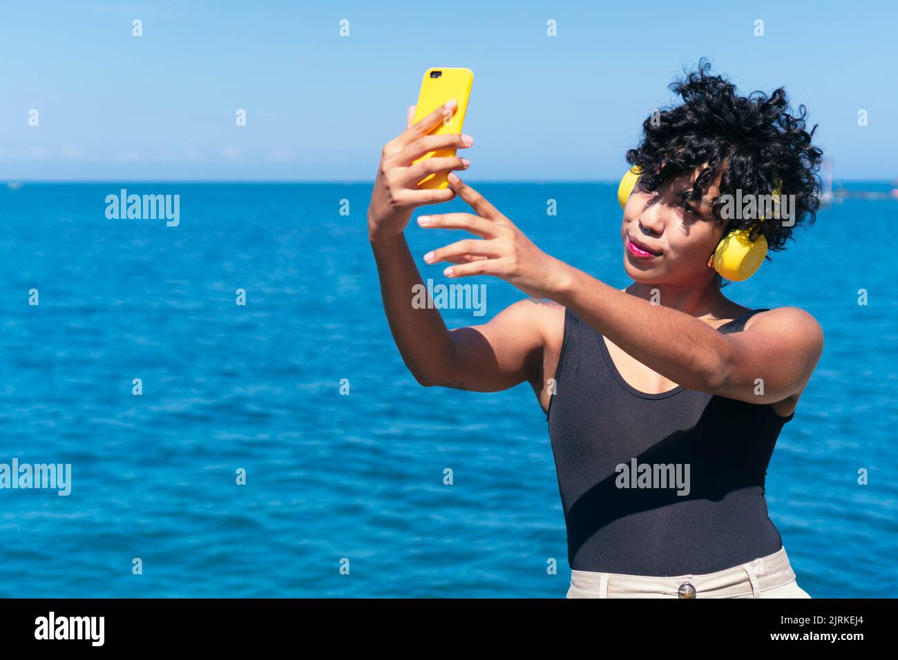 Donna afroamericana con capelli ricci in cuffie wireless che si autoritratto sul cellulare contro il mare alla luce del sole Foto Stock