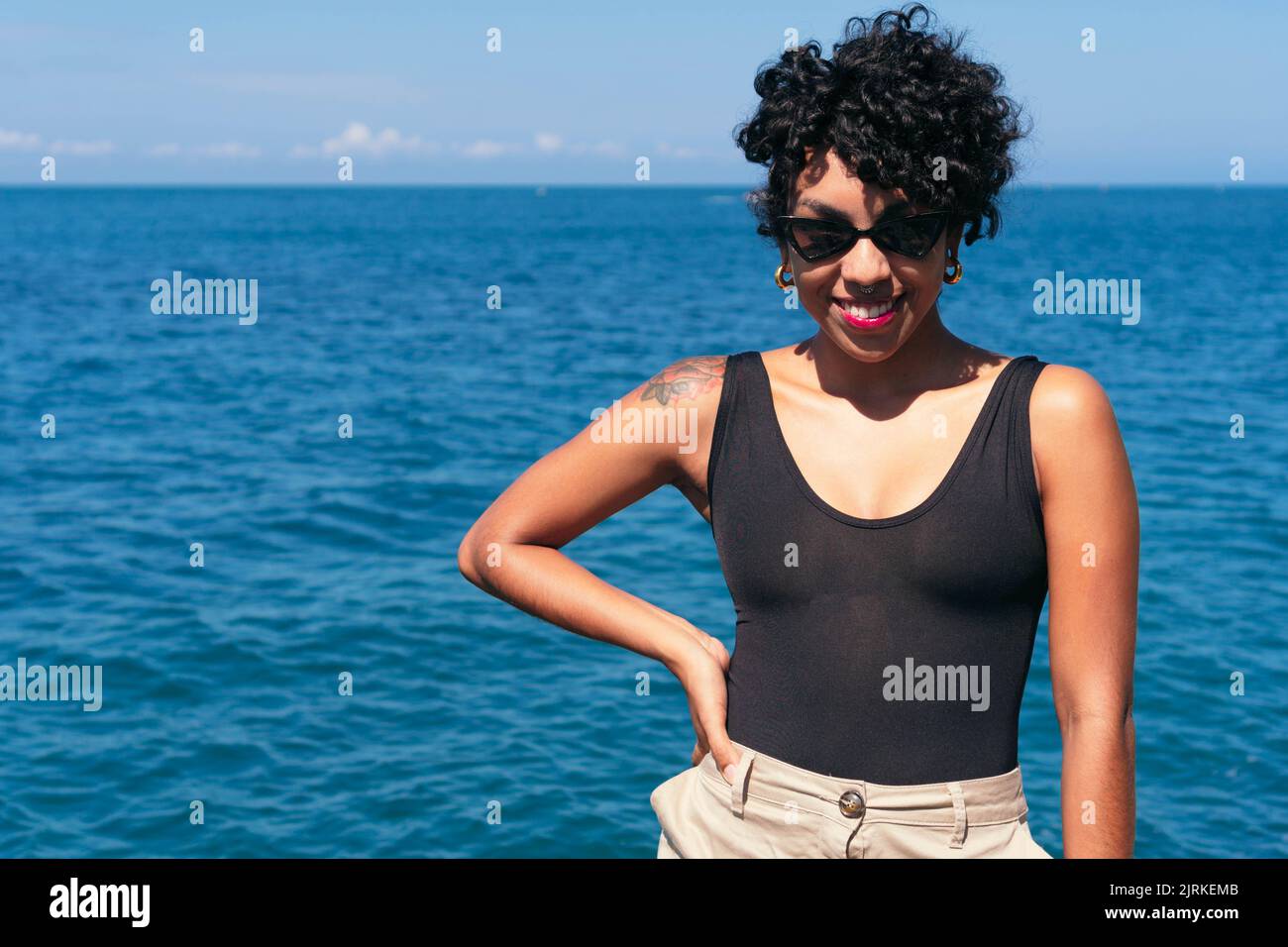 Allegra ragazza afroamericana con tatuaggio e capelli ricci in piedi con la mano sulla vita contro l'oceano sotto il cielo blu Foto Stock