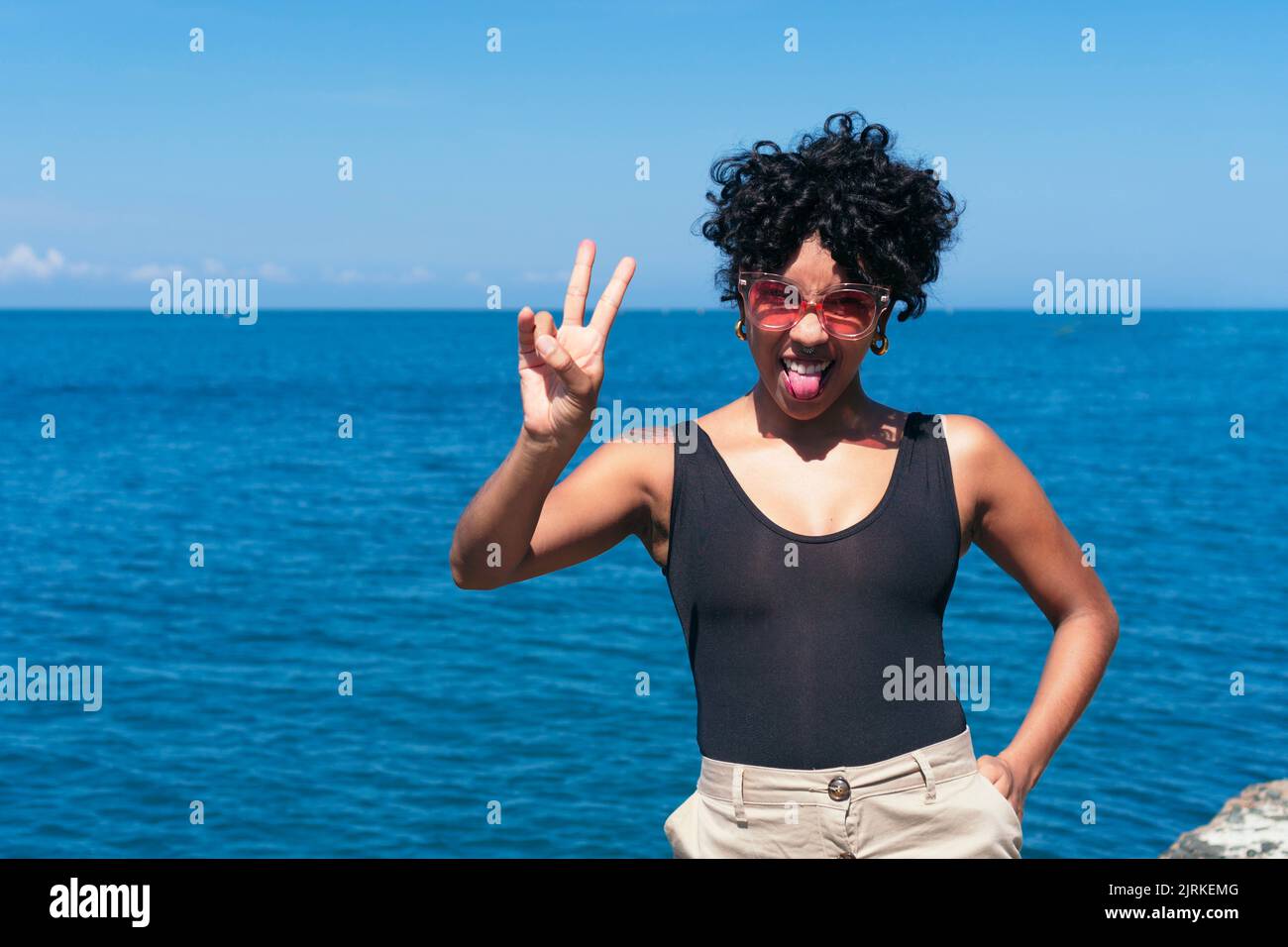 Allegra ragazza afroamericana con tatuaggio e capelli ricci facendo il segno di vittoria mentre si attacca la lingua verso l'oceano sotto il cielo blu Foto Stock