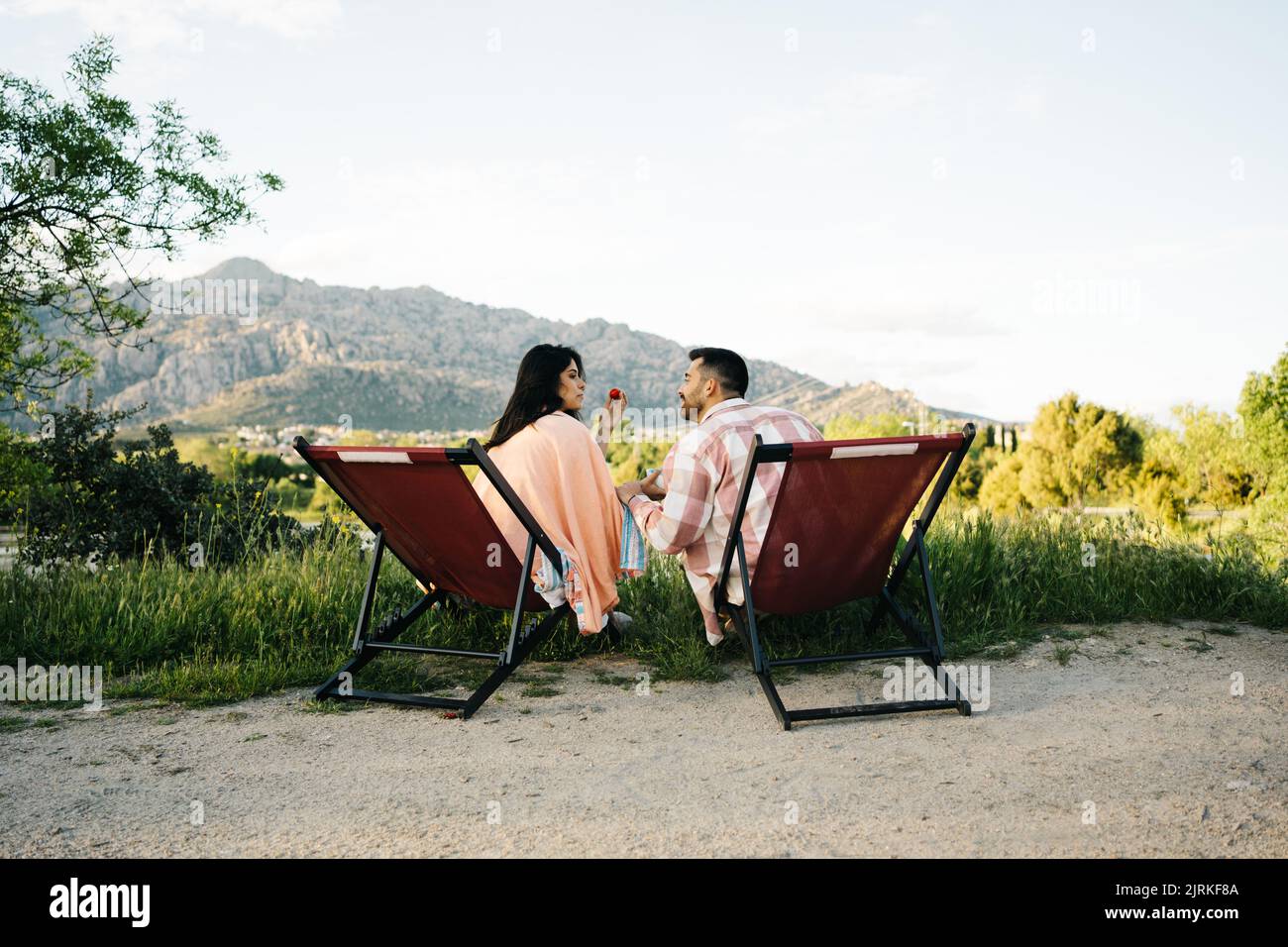 Vista posteriore di una coppia di viaggiatori seduti sulle sedie da campeggio e mangiando fragole mentre si gode un viaggio in montagna Foto Stock