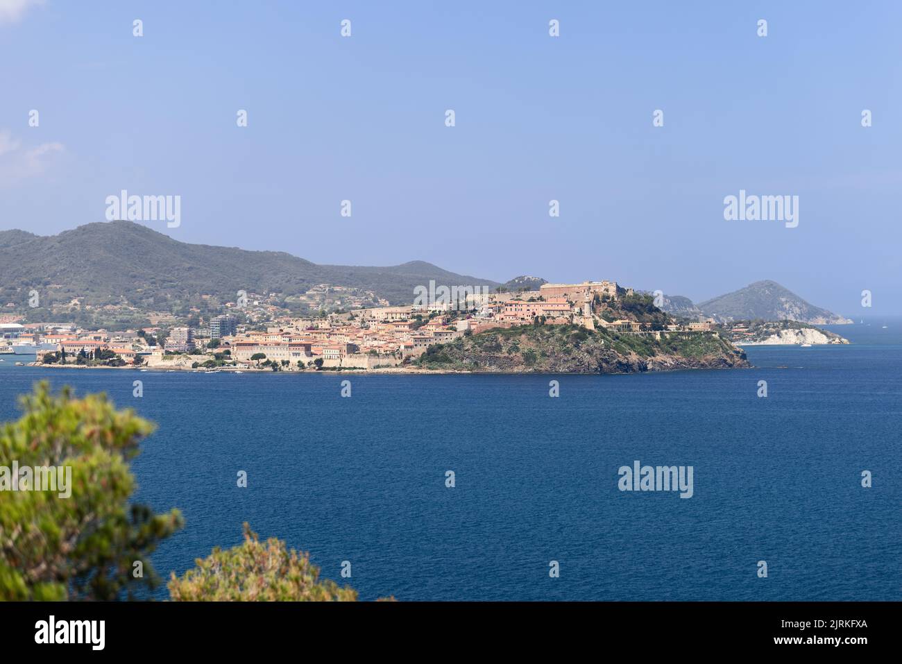 Portoferraio si estende su una penisola fortificata sopra il mare blu dell'isola d'Elba, in Toscana, Italia, con colline verdi sullo sfondo e arbusti costieri in primo piano. Foto Stock