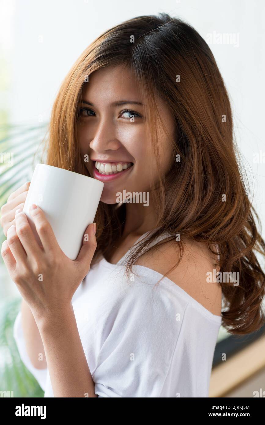 Ritratto in vita di attraente donna vietnamita in posa per la fotografia con un sorriso affascinante mentre si gode caffè fragrante in camera da letto accogliente Foto Stock