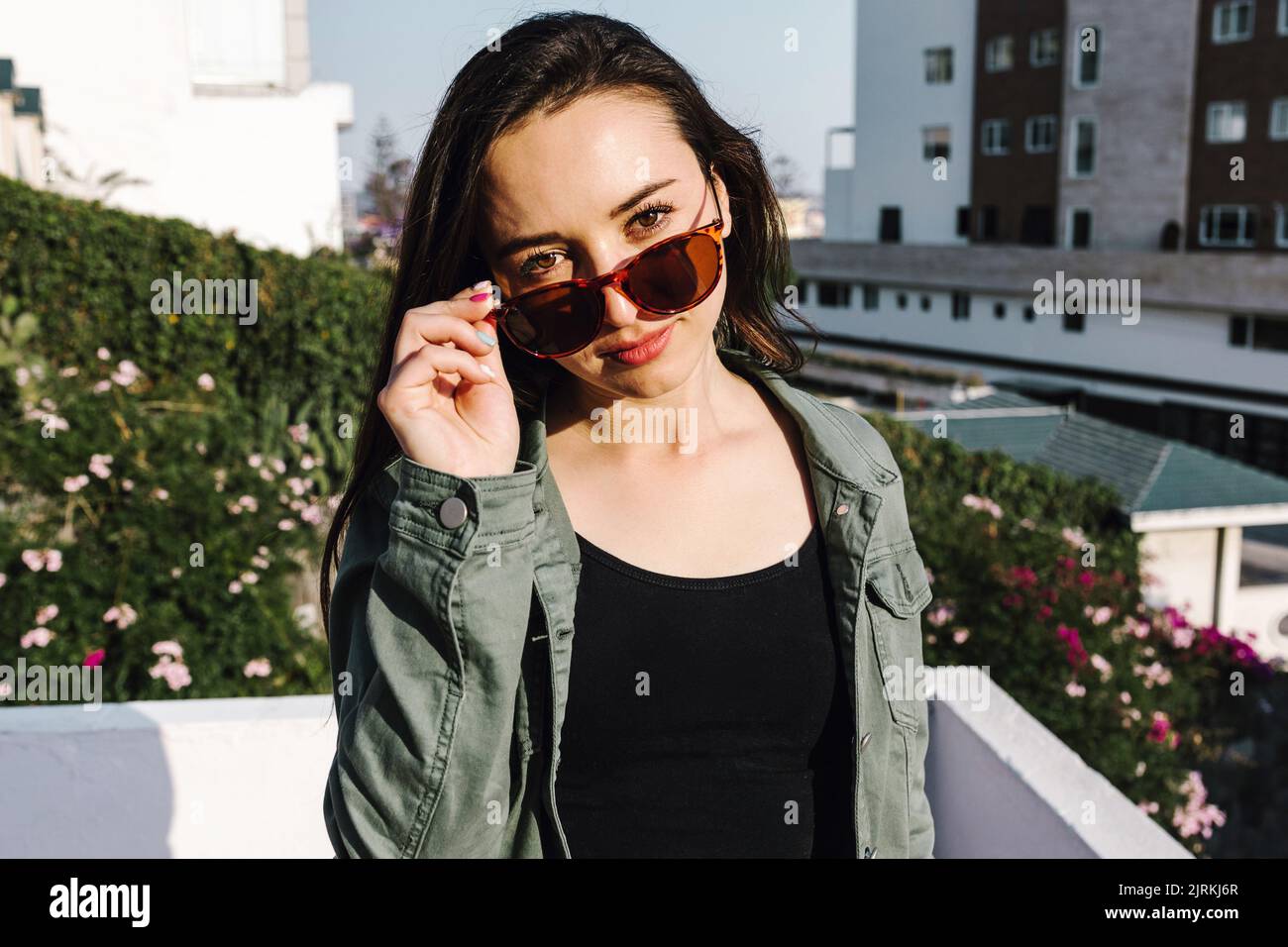 Giovane donna latino-americana in giacca denim e occhiali da sole moderni guardando la macchina fotografica sul balcone contro gli edifici della città alla luce del sole Foto Stock