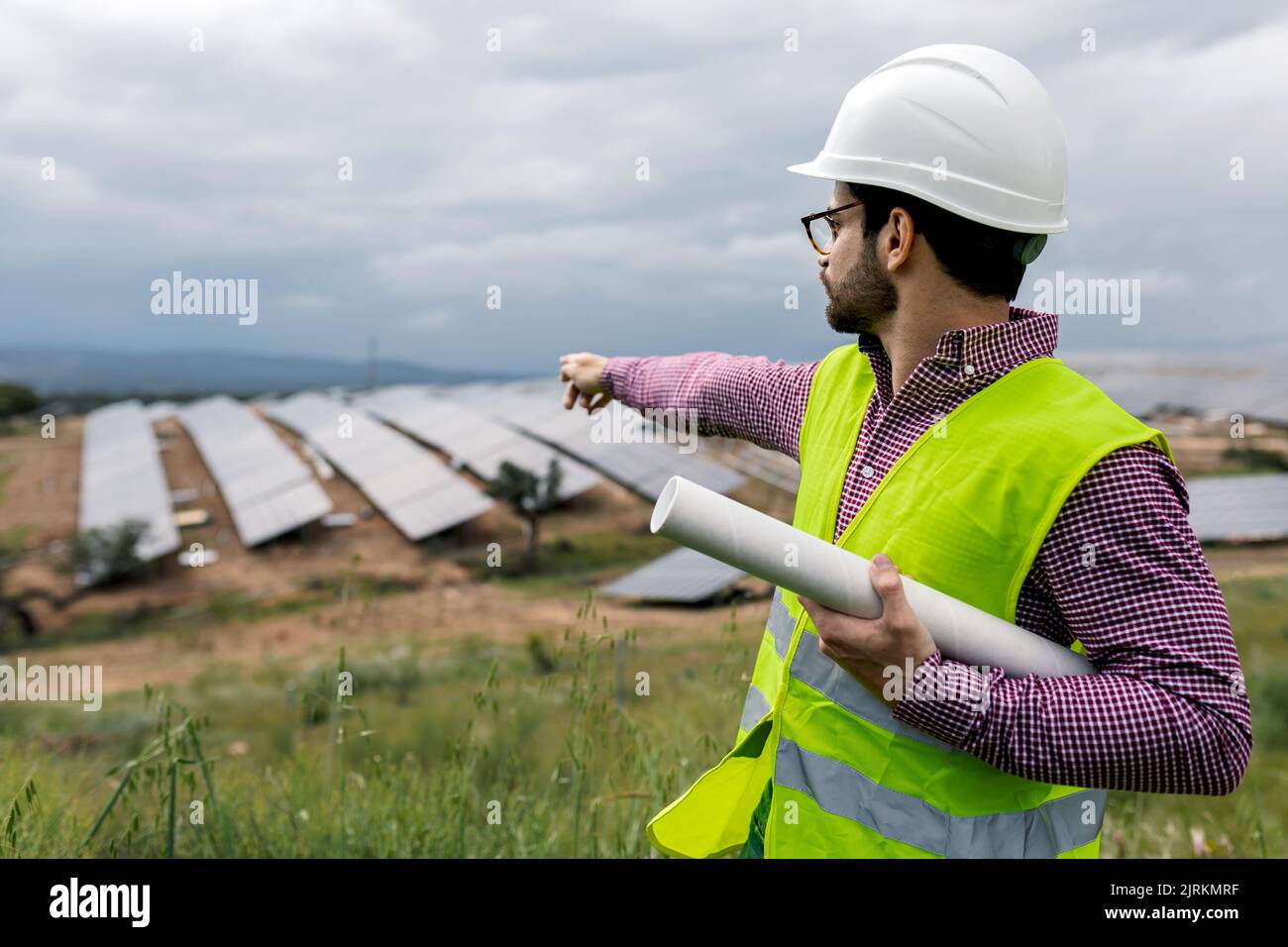 Uomo in elmetto che indica i pannelli fotovoltaici mentre lavora sul sito della centrale di energia solare sullo smartphone Foto Stock