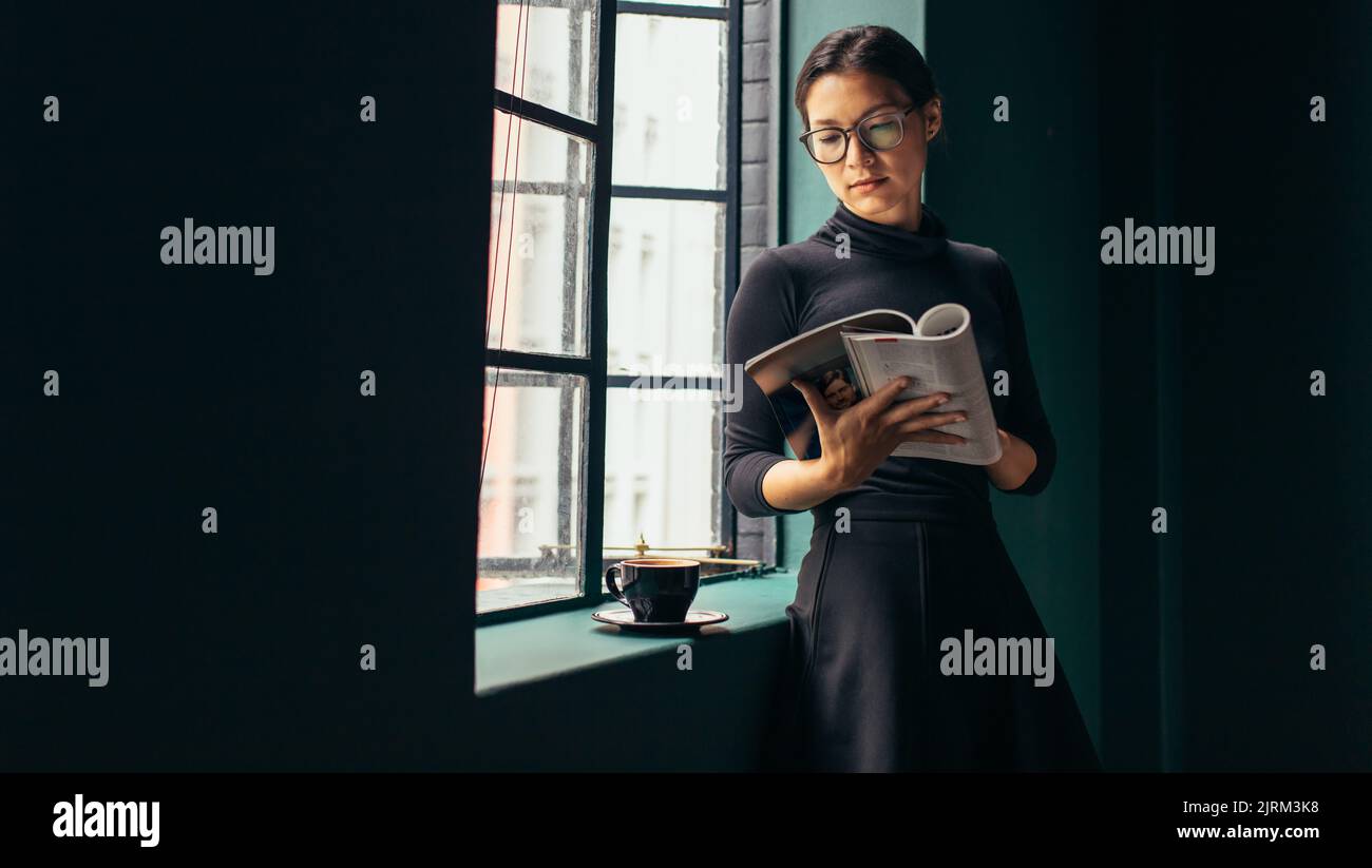 Donna asiatica in piedi accanto alla finestra e libro di lettura. Bella donna lettura rivista di moda. Foto Stock