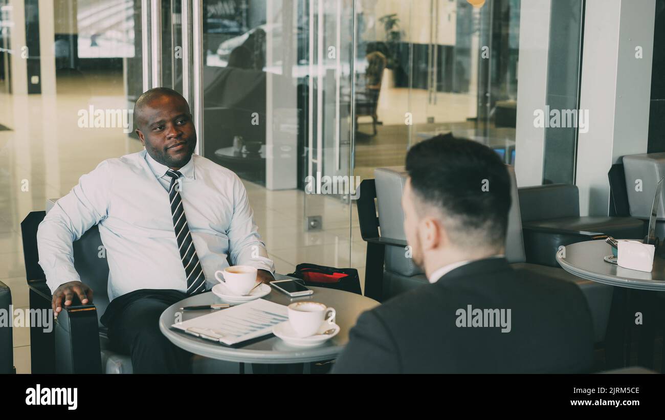 Sorridente uomo d'affari bere caffè e discutere rapporto finanziario di startup con il suo partner in moderno caffè Foto Stock