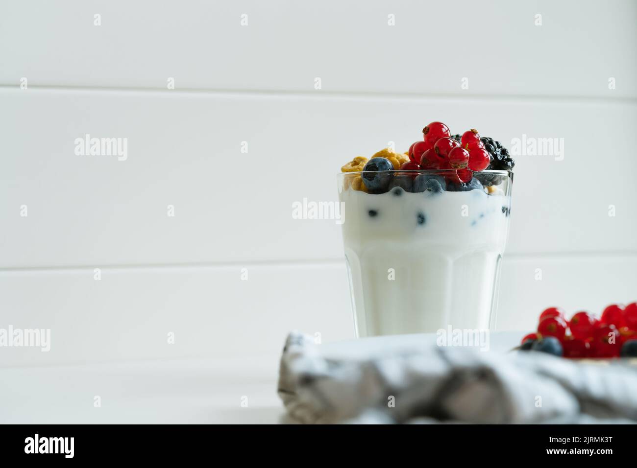 Yogurt, cereali d'avena per la colazione e frutti freschi d'estate in bicchiere su un tavolo bianco. Ribes rossi, mirtilli, more su piatto bianco, Foto Stock