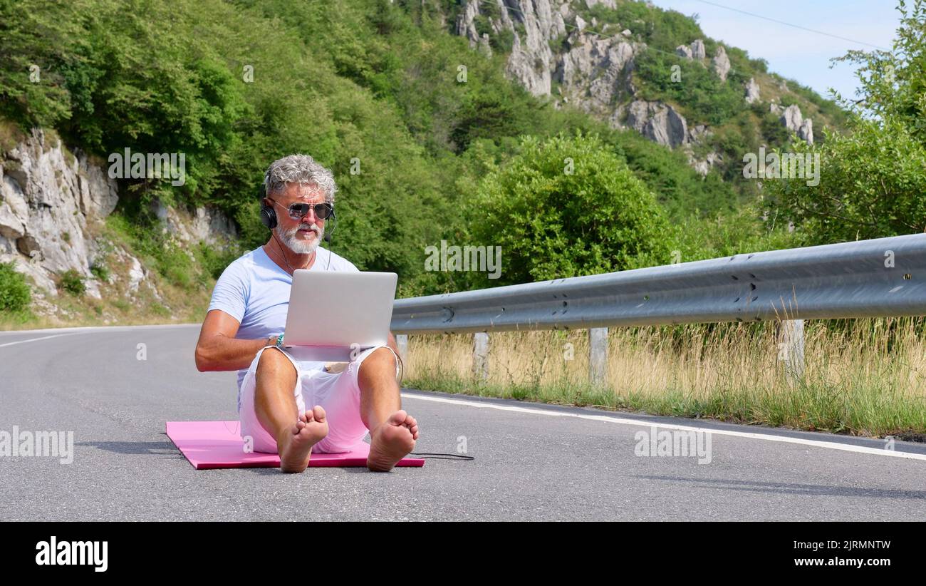 Uomo anziano dai capelli grigi freelancer con barba in occhiali da sole, lavorando su un computer portatile sulla natura, strada, montagna. Un vecchio pazzo e straordinario siede su un tappetino yoga in estate Foto Stock