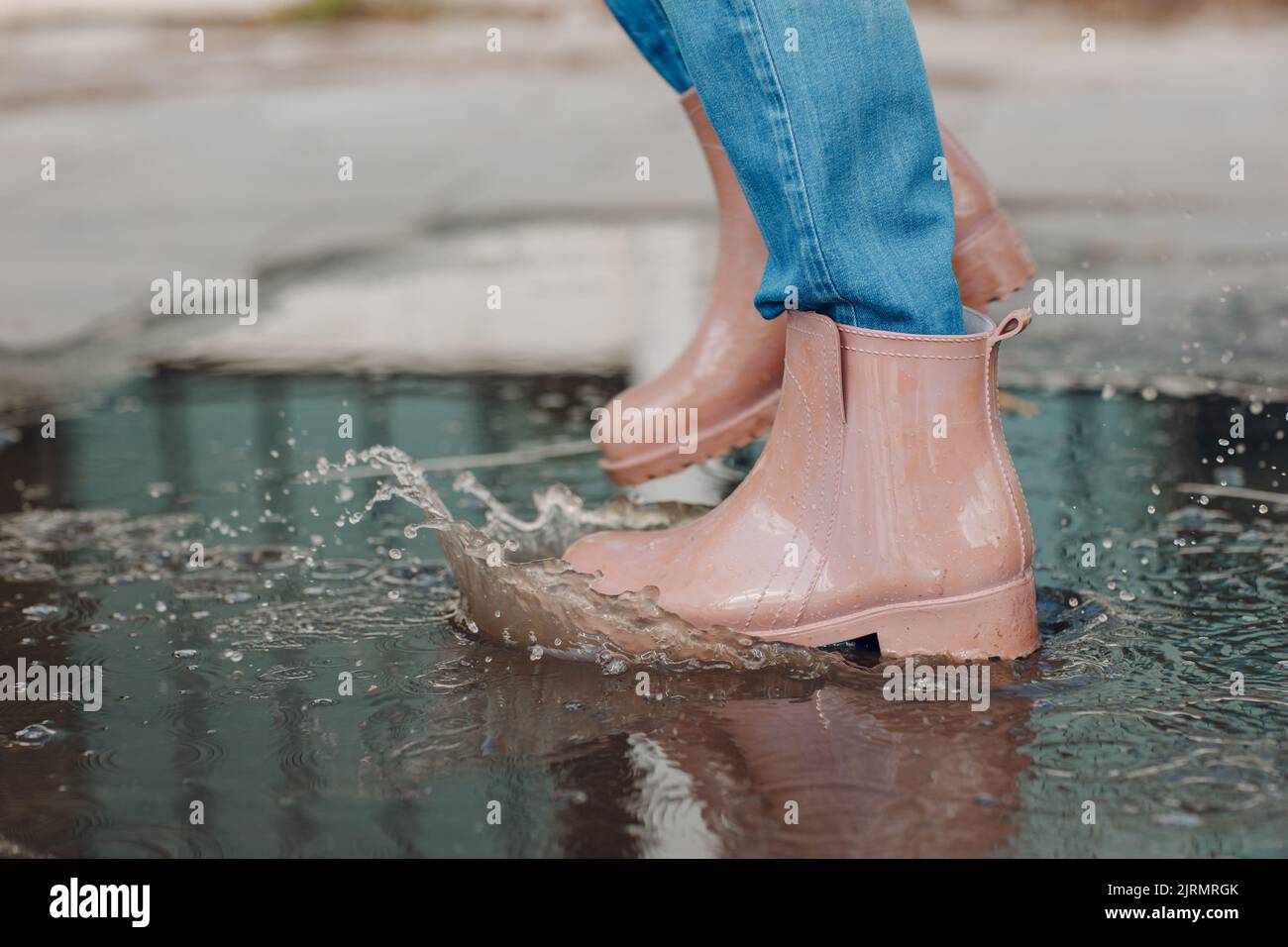 Donna che indossa stivali di gomma da pioggia camminando e saltando nella pozzanghera con spruzzi d'acqua e gocce sotto la pioggia d'autunno. Foto Stock