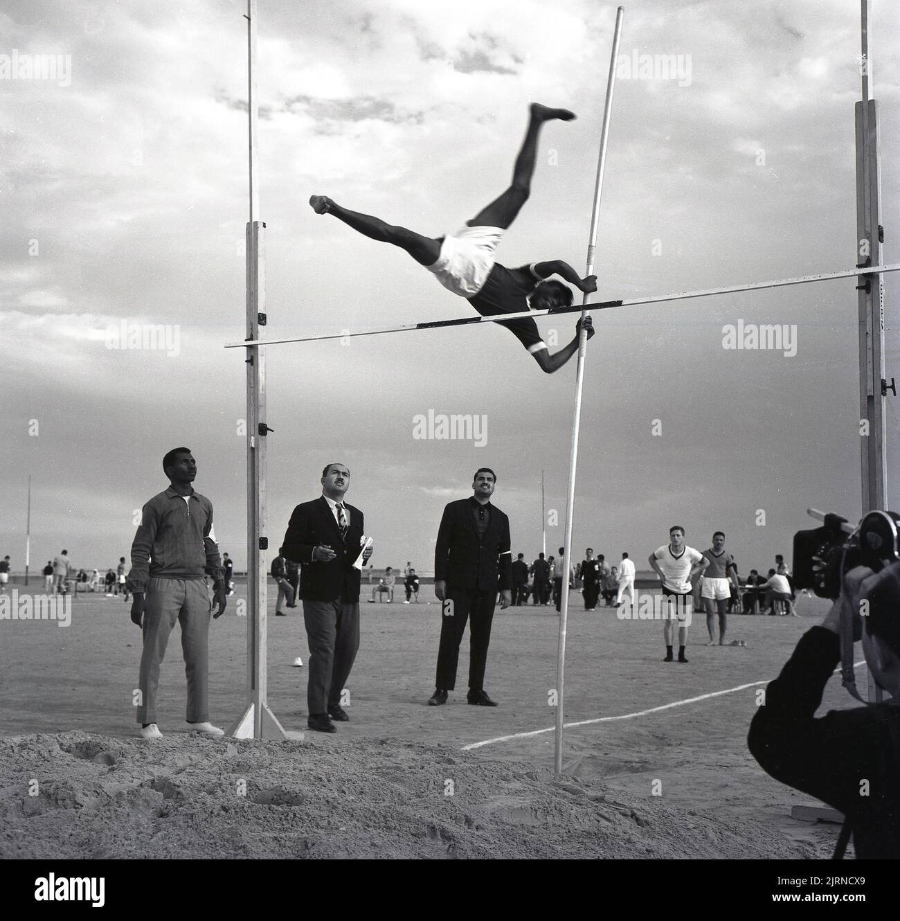 1960s, storico, un cameraman che filma un atleta arabo maschio, un polevaulter, con la sua posizione non convenzionale dei piedi, mentre tenta di liberare il bar, sostenuto su due montanti, Jeddah, Arabia Saudita. Uno dei principali eventi di salto in atletica, è stato un evento di medaglia completa alle Olimpiadi dal 1896. Foto Stock