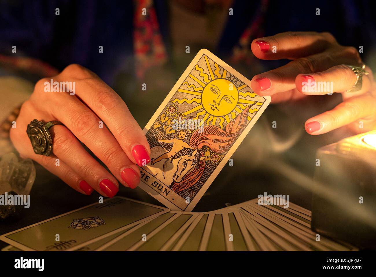 Fortune Teller mani femminili che mostrano la carta del tarocco del Sole, simbolo di positività e ottimismo, durante una lettura. Primo piano con luce a candela e fumo, moo Foto Stock