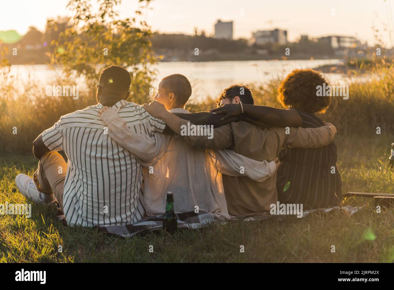 Vista posteriore di buoni vecchi amici multiculturali abbracciare mentre si siede su una coperta sull'erba, avendo un picnic di fine settimana nella natura vicino al fiume, parlando, bevendo la birra e ammirando la vista. Ora del tramonto. Foto di alta qualità Foto Stock