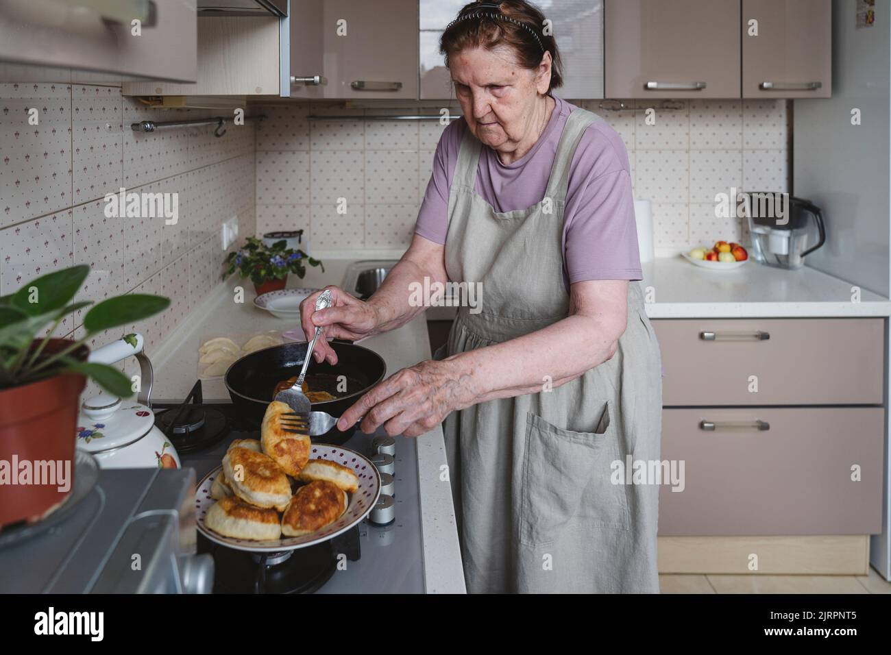 Una donna anziana frigge torte in cucina. Foto Stock