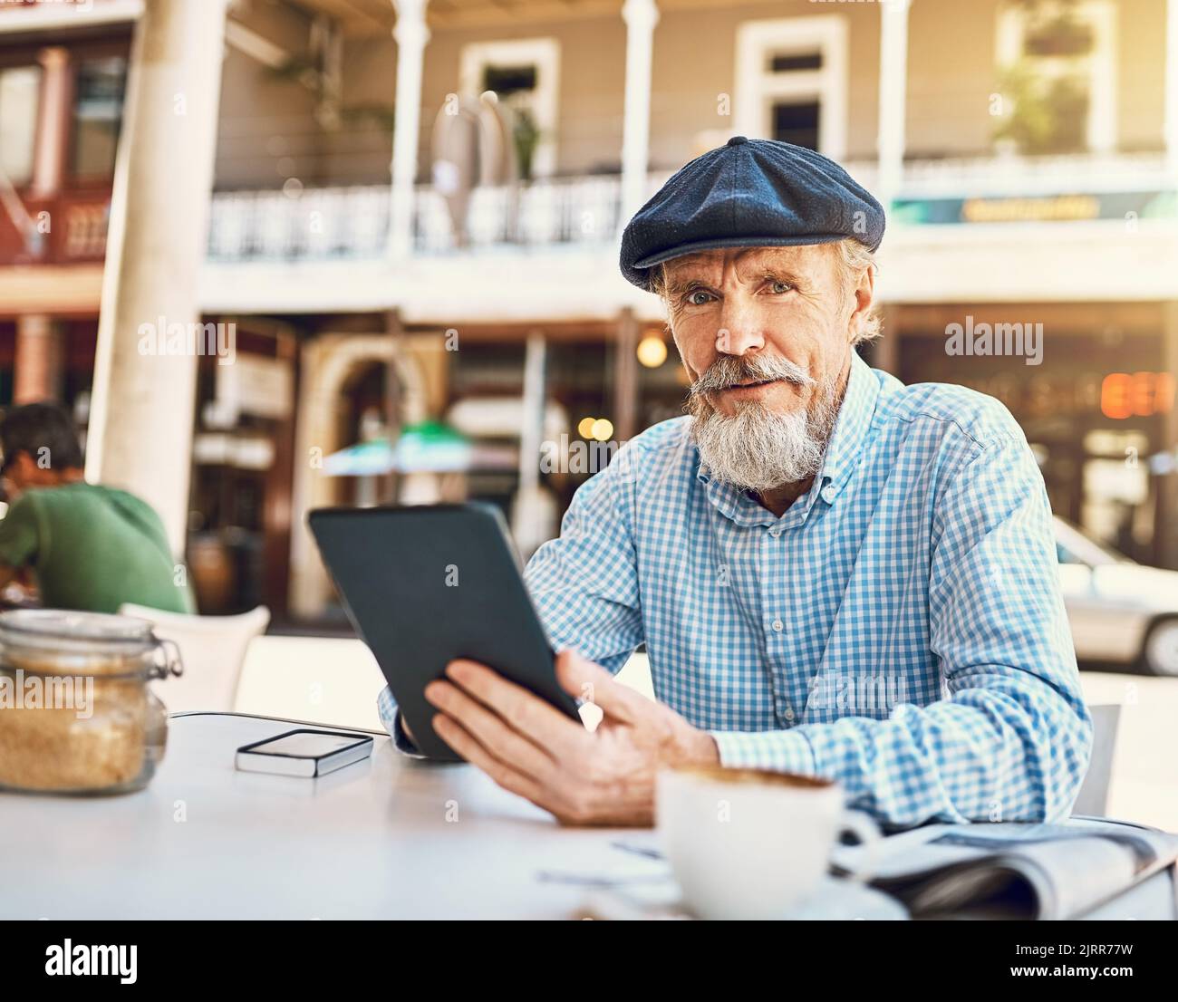 Mi limiterò a stare al passo con i miei follower sui social media. Ritratto di un uomo anziano felice che usa il suo tablet mentre si siede in un bar sul marciapiede. Foto Stock