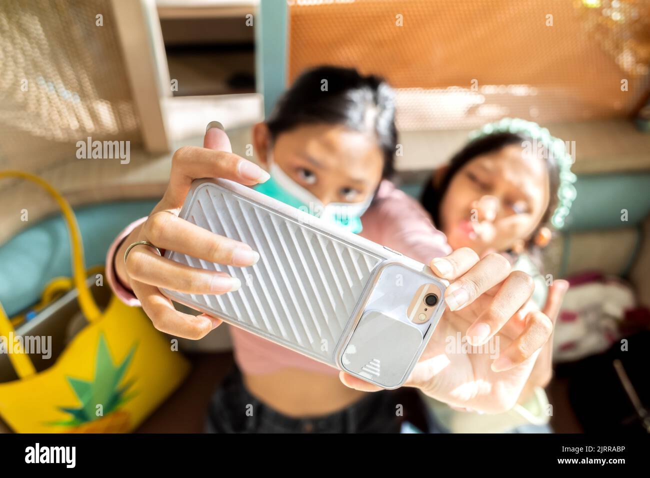 Ragazza adolescente del sud-est asiatico indossare maschera fare selfie insieme con sorella utilizzando smartphone in un ristorante Foto Stock