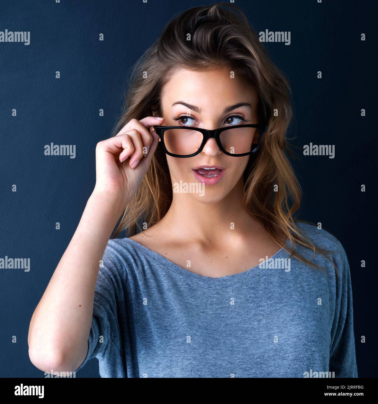 È questo che penso sia. Studio shot di una ragazza adolescente carina in occhiali che alzano le sopracciglia a sorpresa posando su uno sfondo scuro. Foto Stock