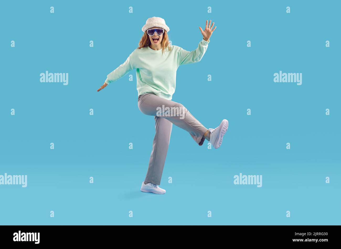 Donna divertente in felpa, pantaloni, cappello da secchio e occhiali da sole che ballano su sfondo blu studio Foto Stock
