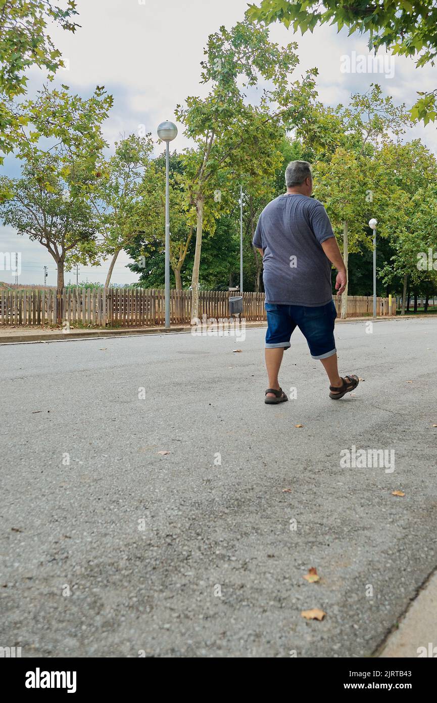 Obese che cammina in un parco all'aperto circondato da alberi con lampioni e una recinzione. Foto Stock