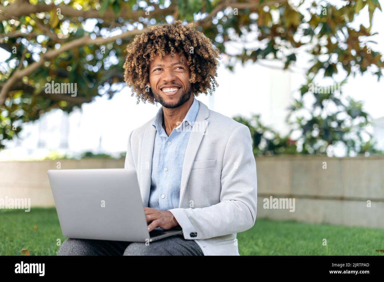 Bello attraente gara mista riuscito uomo d'affari, manager, freelancer, elegantemente vestito, seduto all'aperto con un computer portatile, guardando al lato, sognando, sorridendo amichevole Foto Stock