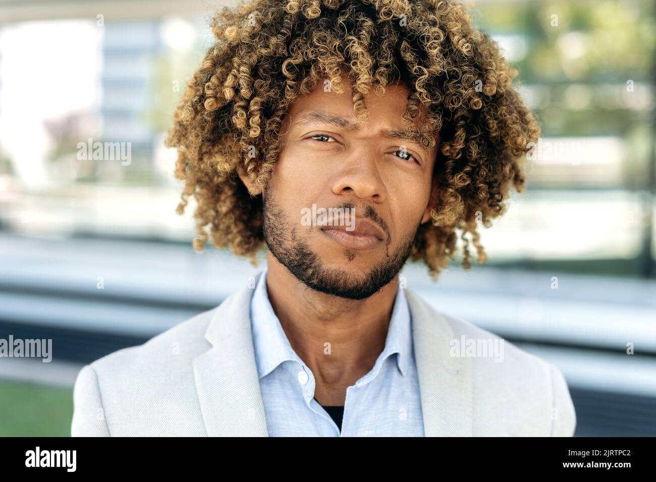 Primo piano di un orgoglioso sicuro serio elegante capelli ricci brasiliano o ispano maschio, imprenditore di successo, uomo d'affari, in piedi all'aperto, guardando la fotocamera Foto Stock