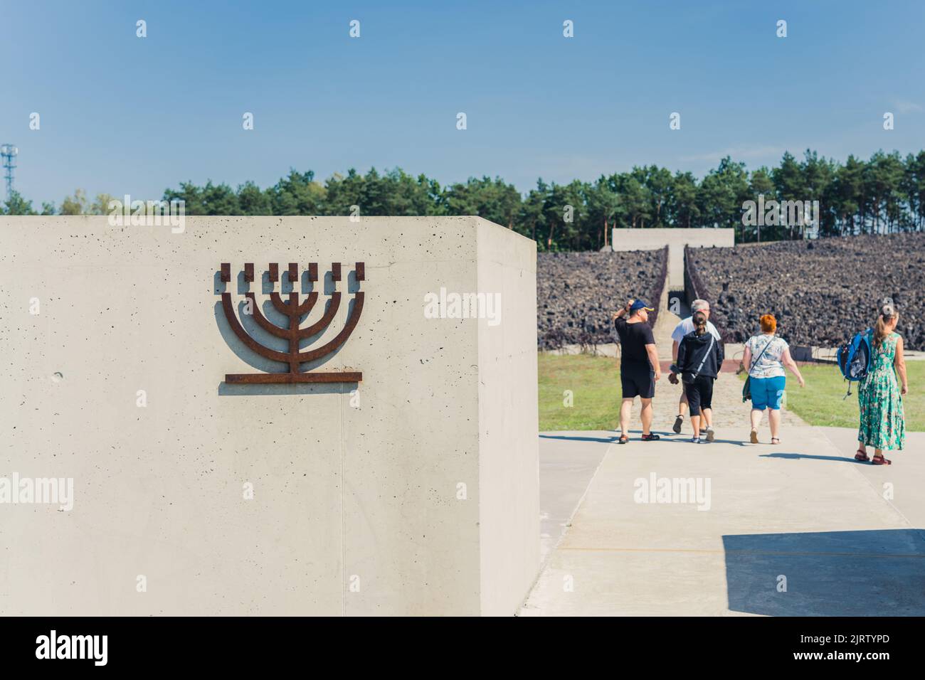 08.27.2022 - Belzec, Polonia - campo di sterminio nazista di Belzec. Breve muro di conrete con simbolo di menora metallica di fronte all'ingresso dell'ex campo di sterminio nazista. Turisti al memoriale dell'Olocausto. Scatto orizzontale. Foto di alta qualità Foto Stock