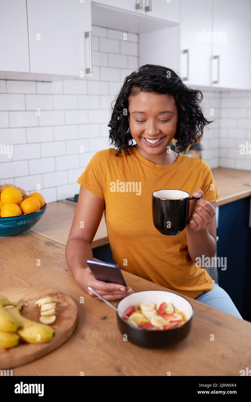 Una giovane donna multietnica beve caffè e guarda il suo telefono sopra la colazione Foto Stock