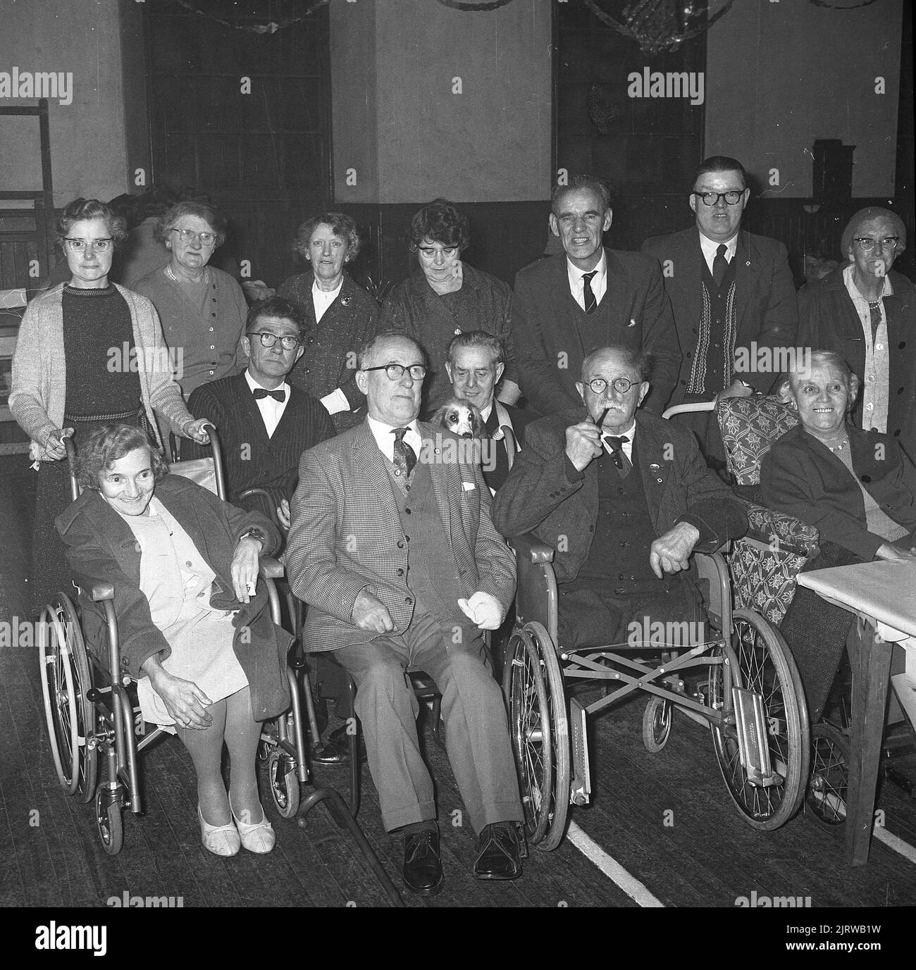 1965, storico, Red Cross tè party per disabili, foto di gruppo che mostra volontari in piedi con i loro ospiti in sedia a rotelle, Fife, Scozia, Regno Unito. Una delle principali organizzazioni di beneficenza, la Croce Rossa britannica, fornisce sostegno alle persone anziane e vulnerabili, aiutandole a vivere in modo indipendente nelle loro case. L'ente benefico sostiene anche l'NHS, aiutando i pazienti a tornare a casa dall'ospedale, facilitando il flusso dei pazienti e riducendo le loro necessità immediate. I principi della Croce Rossa sono: Umanità, imparzialità, neutralità, indipendenza, servizio volontario, unità e universalità. Foto Stock