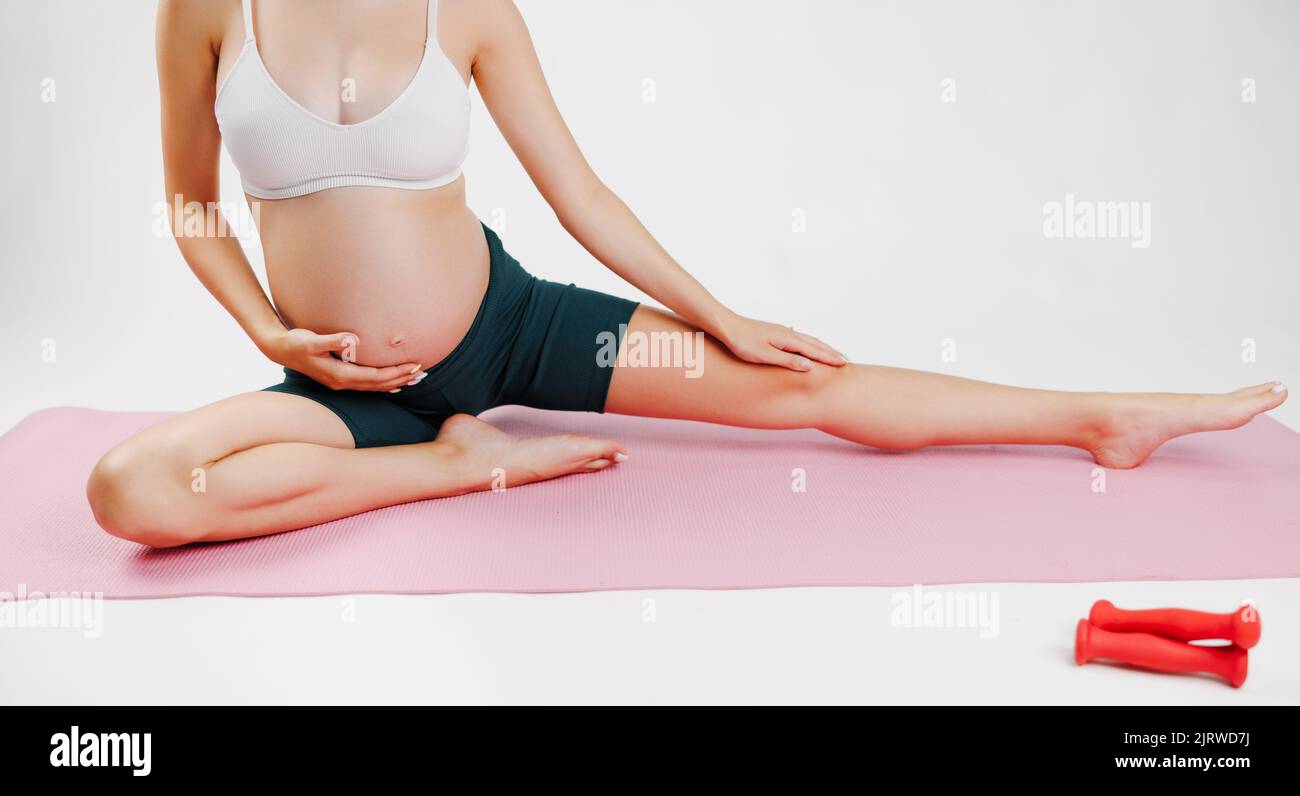 Ritratto di una adorabile giovane donna incinta sportiva in abbigliamento sportivo alla moda che allunga la gamba sinistra, isolata su una parete bianca illuminata dal sole. Allenamento, yoga e. Foto Stock