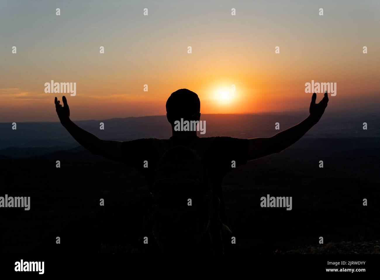 Tramonto con una silhouette di un giovane con un cappello in un atteggiamento di gratitudine al sole, con uno sfondo di montagne. Foto Stock