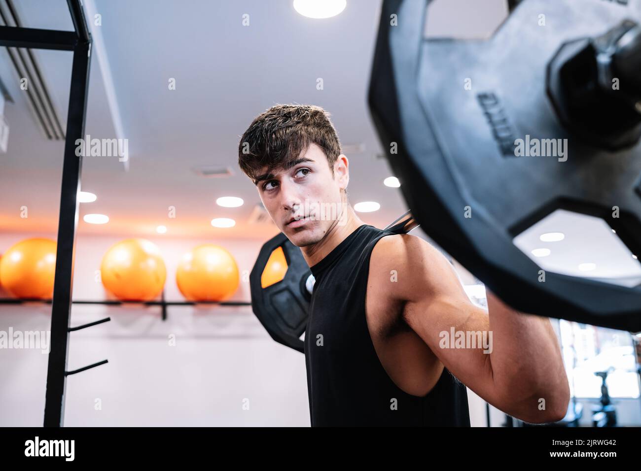 Sportivo giovane e forte con il barbell sulle spalle che guarda lontano durante l'allenamento di sollevamento pesi durante il giorno in palestra Foto Stock