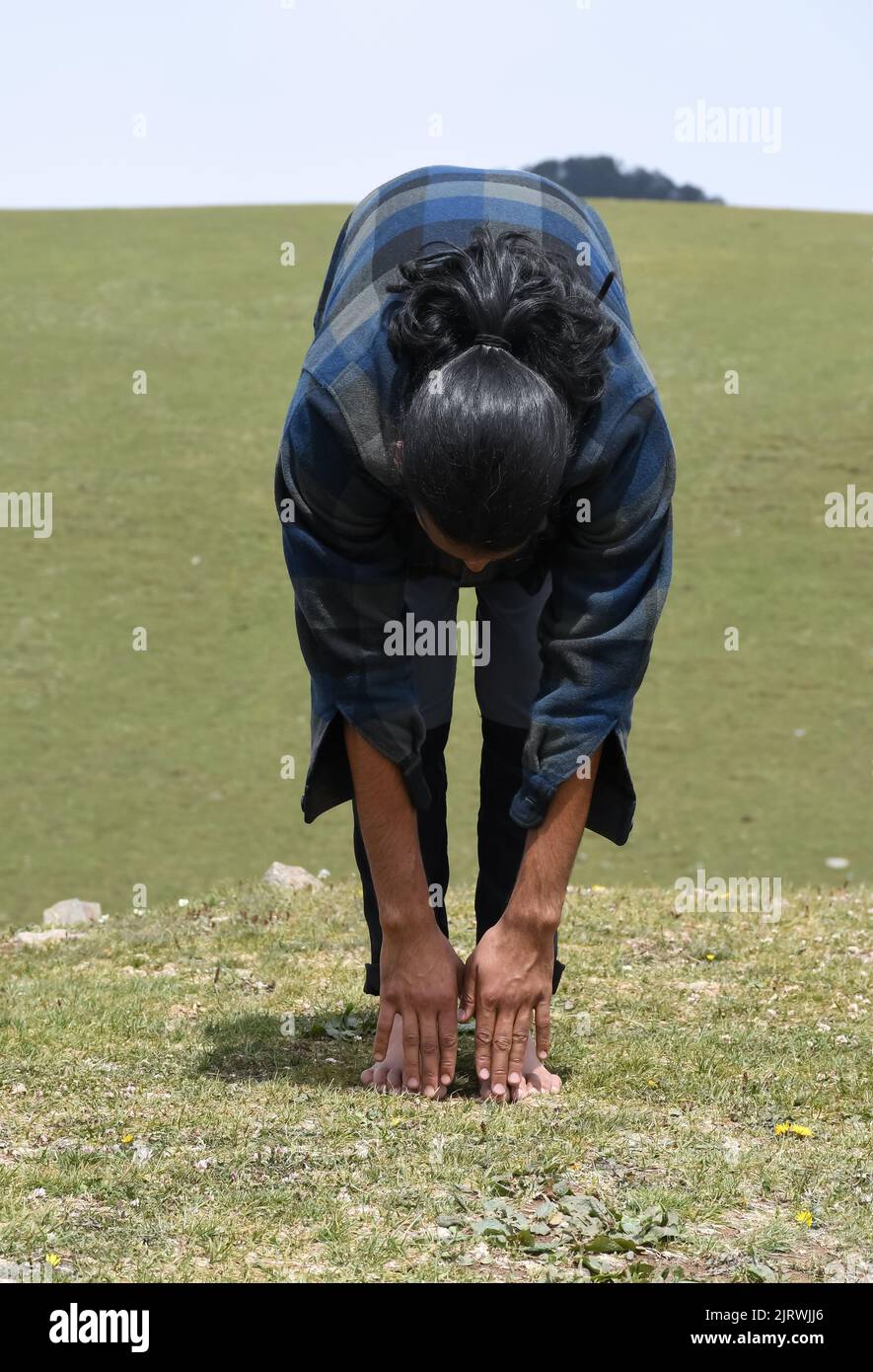 Un giovane uomo dai capelli lunghi che fa il riscaldamento da Utttanasana Yoga posa (tocco in piedi della punta) l'esercitazione di stiramento nella natura. Foto Stock