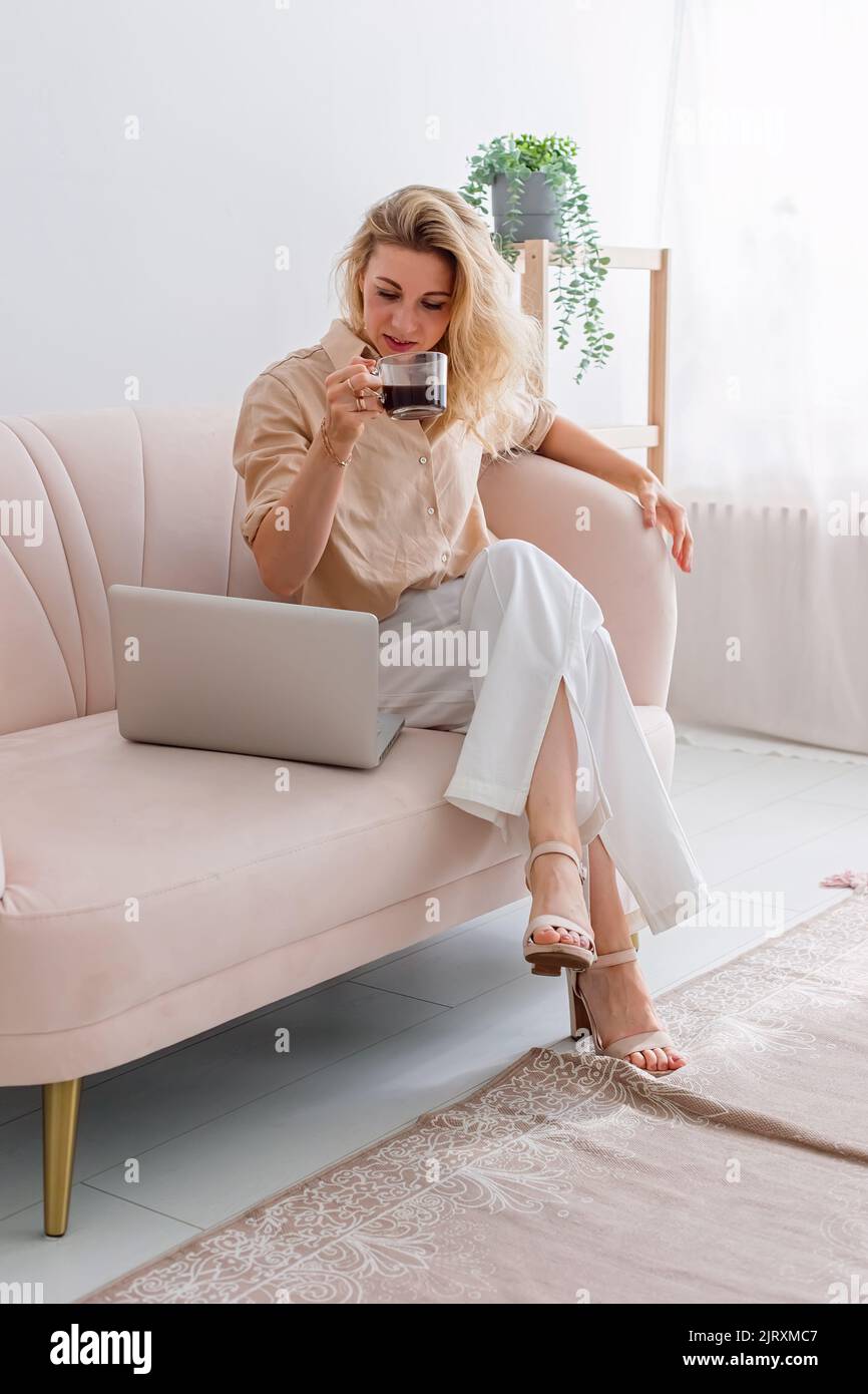 Una donna d'affari bionda, che lavora su un computer portatile, si siede sul divano con coppa COFee Foto Stock