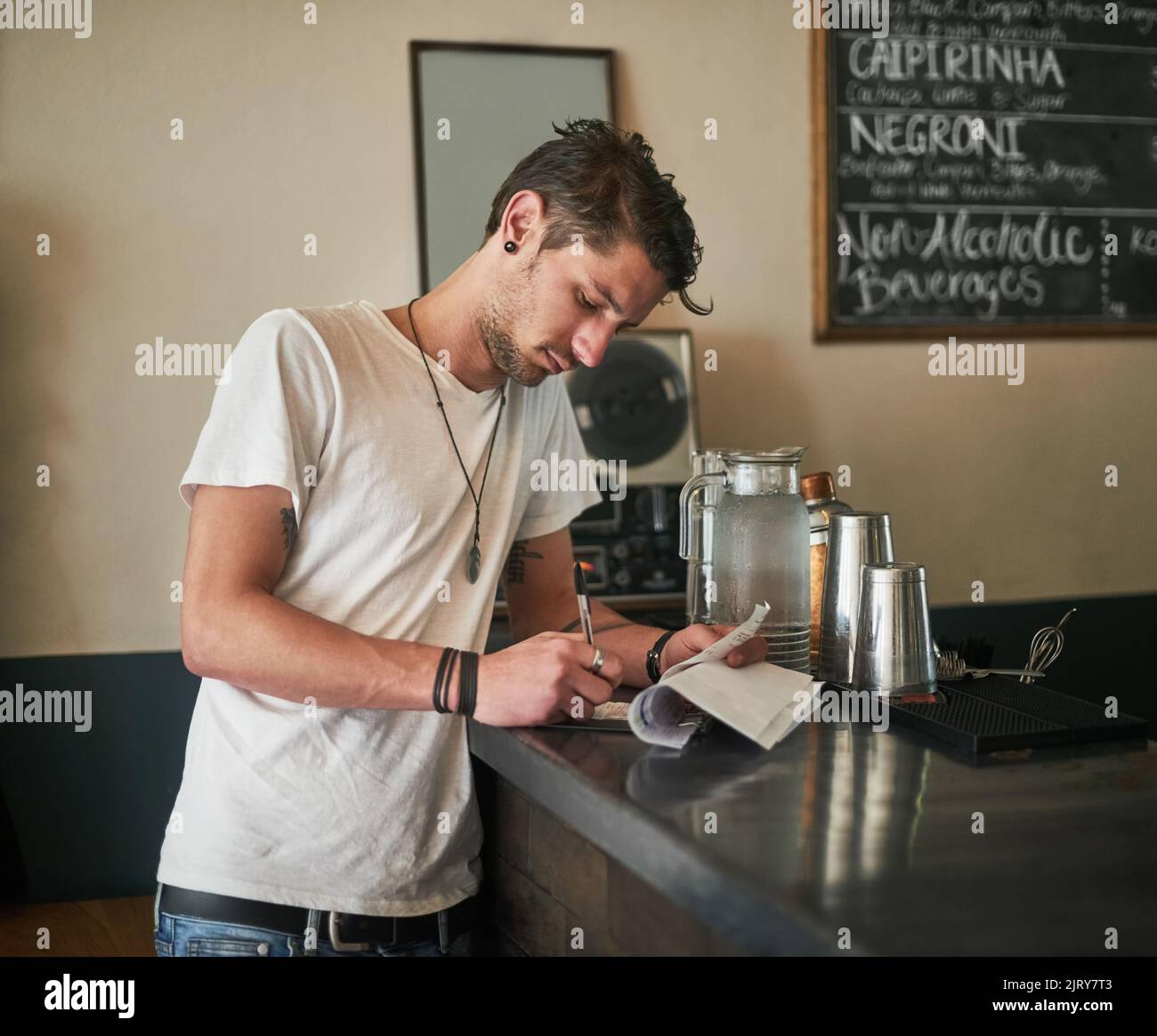 Un altro giorno nella vita di un barkeep. Un barman giovane e focalizzato che prende appunti sulle ricevute dietro il bar. Foto Stock
