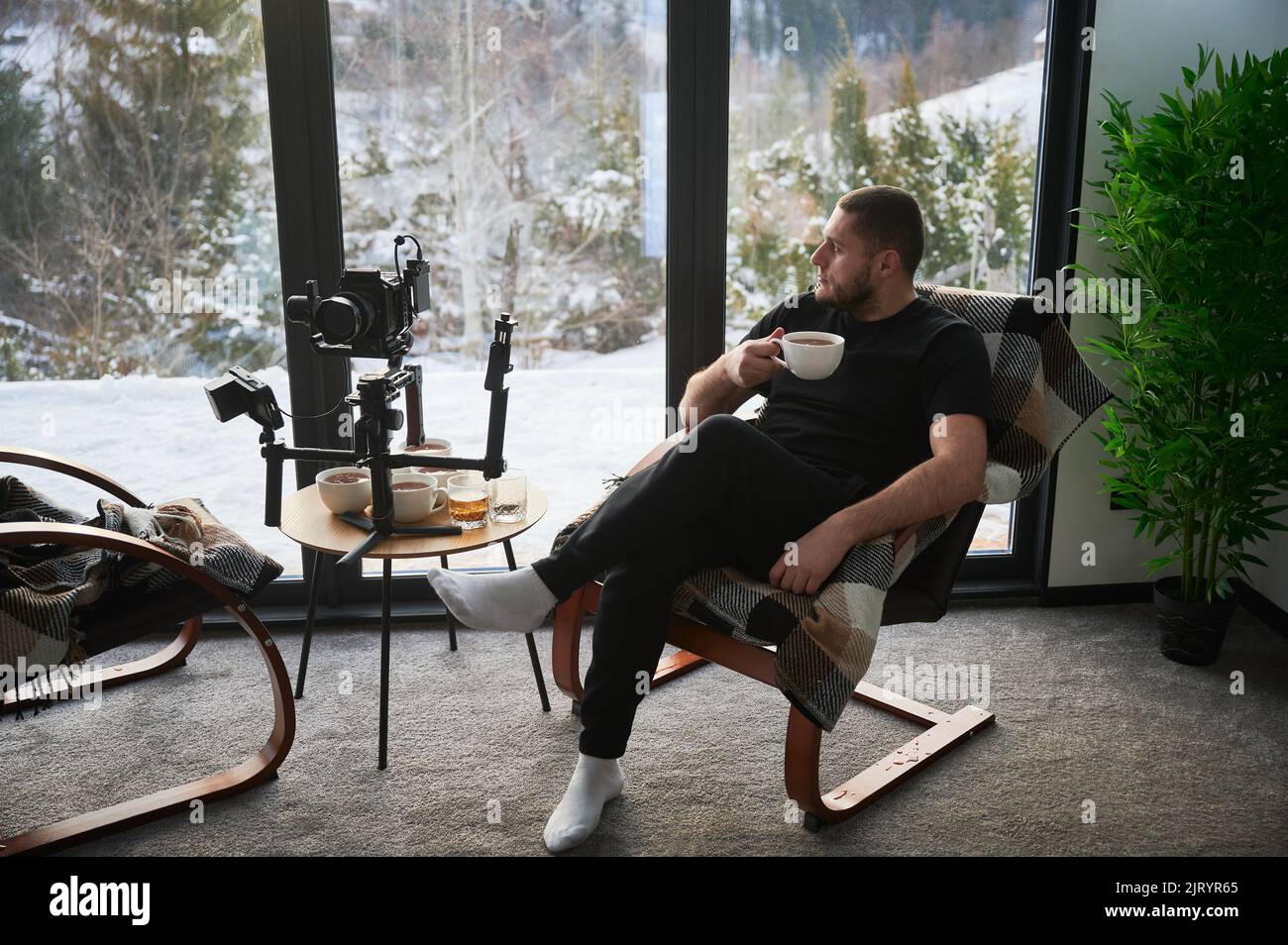 Uomo videografo che riposa sulla poltrona, bevendo il tè vicino alla sua macchina fotografica montata su stabilizzatore gimbal attrezzatura. Foto Stock