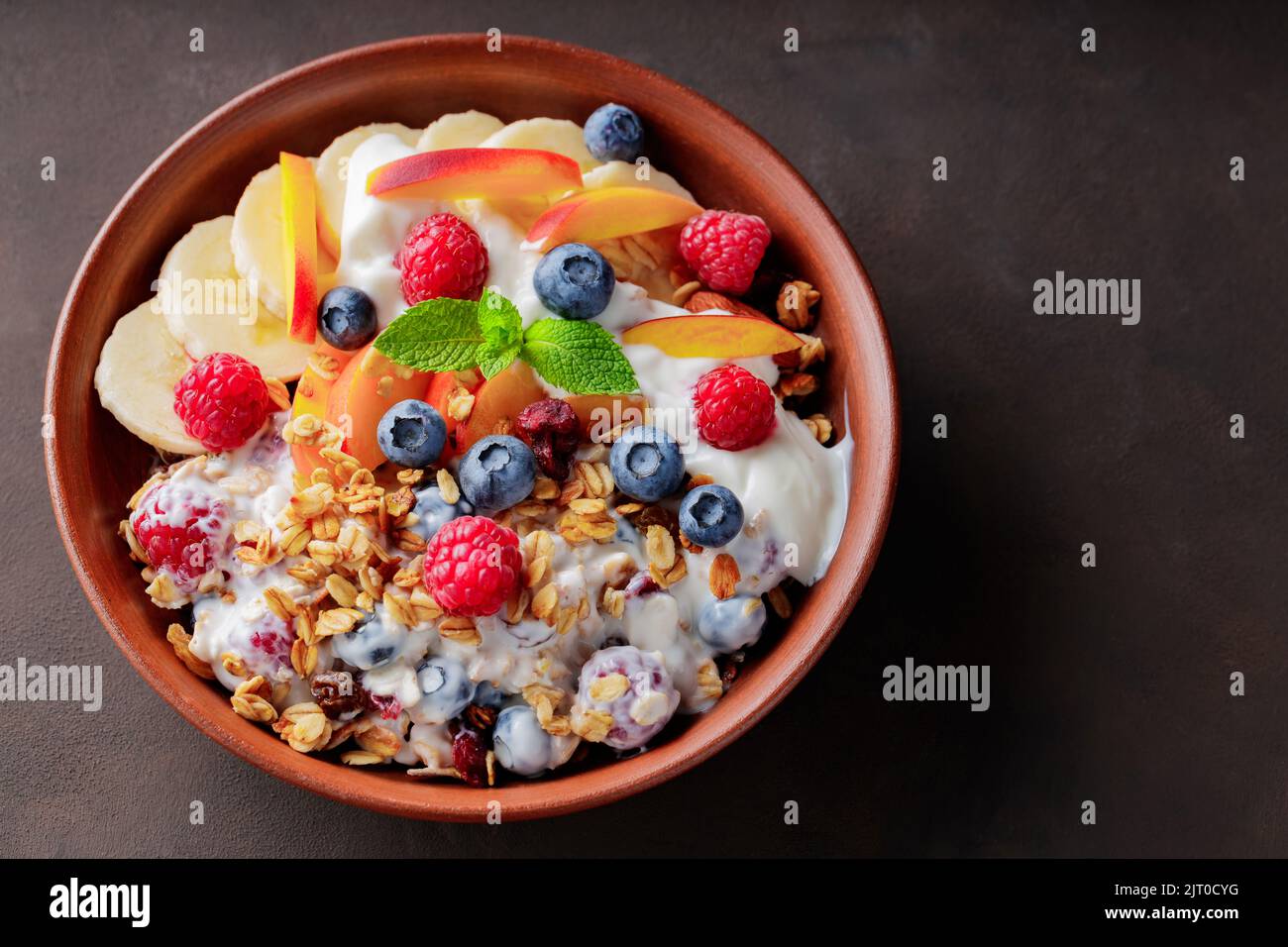 Colazione granola con yogurt, mirtillo, lampone, banana e pesca. Colazione sana con yogurt, granola al forno e frutta su un piatto di argilla. Vista superiore Foto Stock