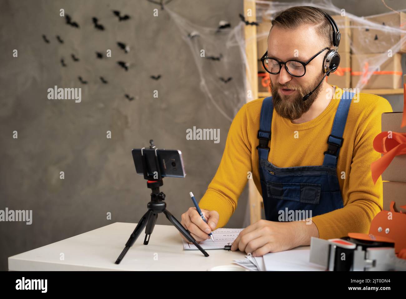 barbuto uomo in vetri imprenditore si siede in halloween decorato home office. Scrive con una penna su carta. Vendendo le merci in onore della festa di Foto Stock