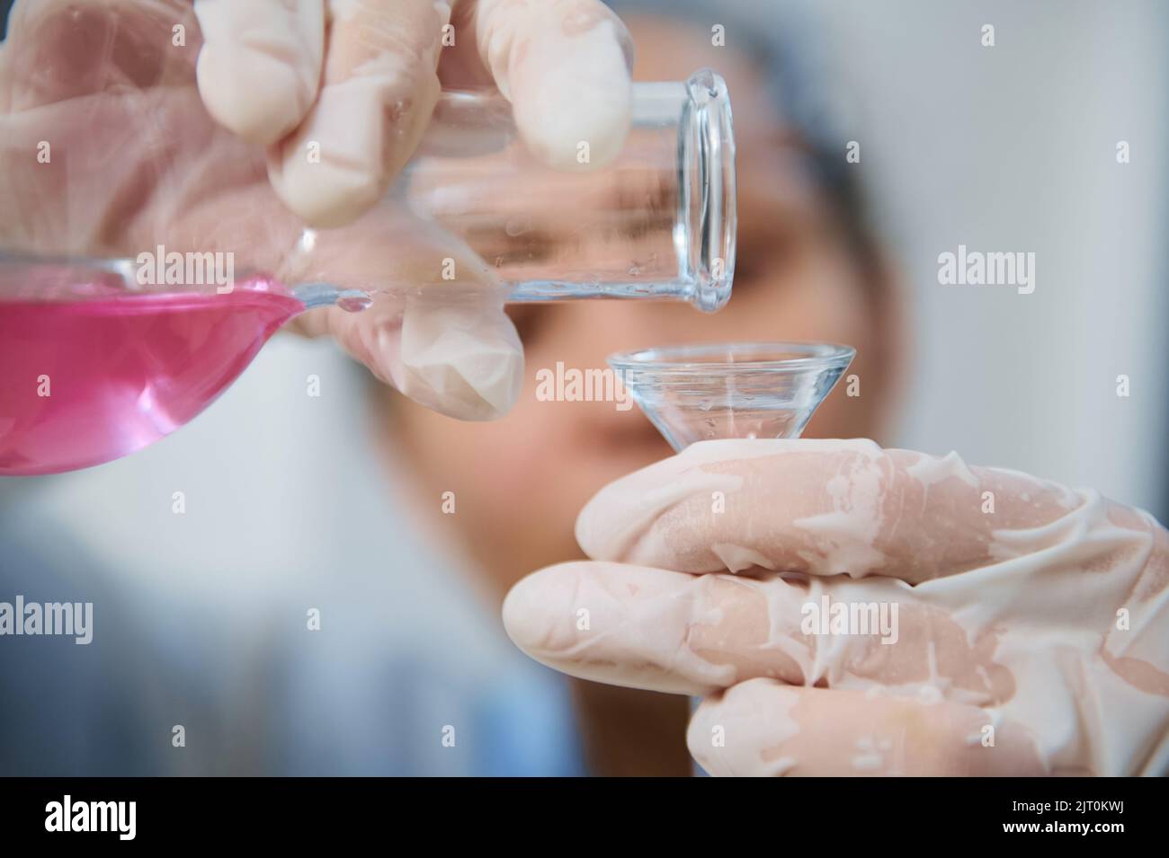 Dettagli: Mani del personale di laboratorio con guanti che versano la sostanza chimica liquida rosa da un pallone di vetro attraverso un imbuto Foto Stock