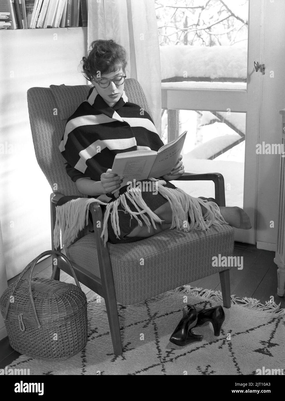 Negli anni '1950s. Una giovane donna a casa si siede in poltrona leggendo un libro. Ha un blanke caldo intorno a lei. È inverno e la neve è visibile attraverso la finestra. Svezia 1953. Rif. Conard 2317 Foto Stock