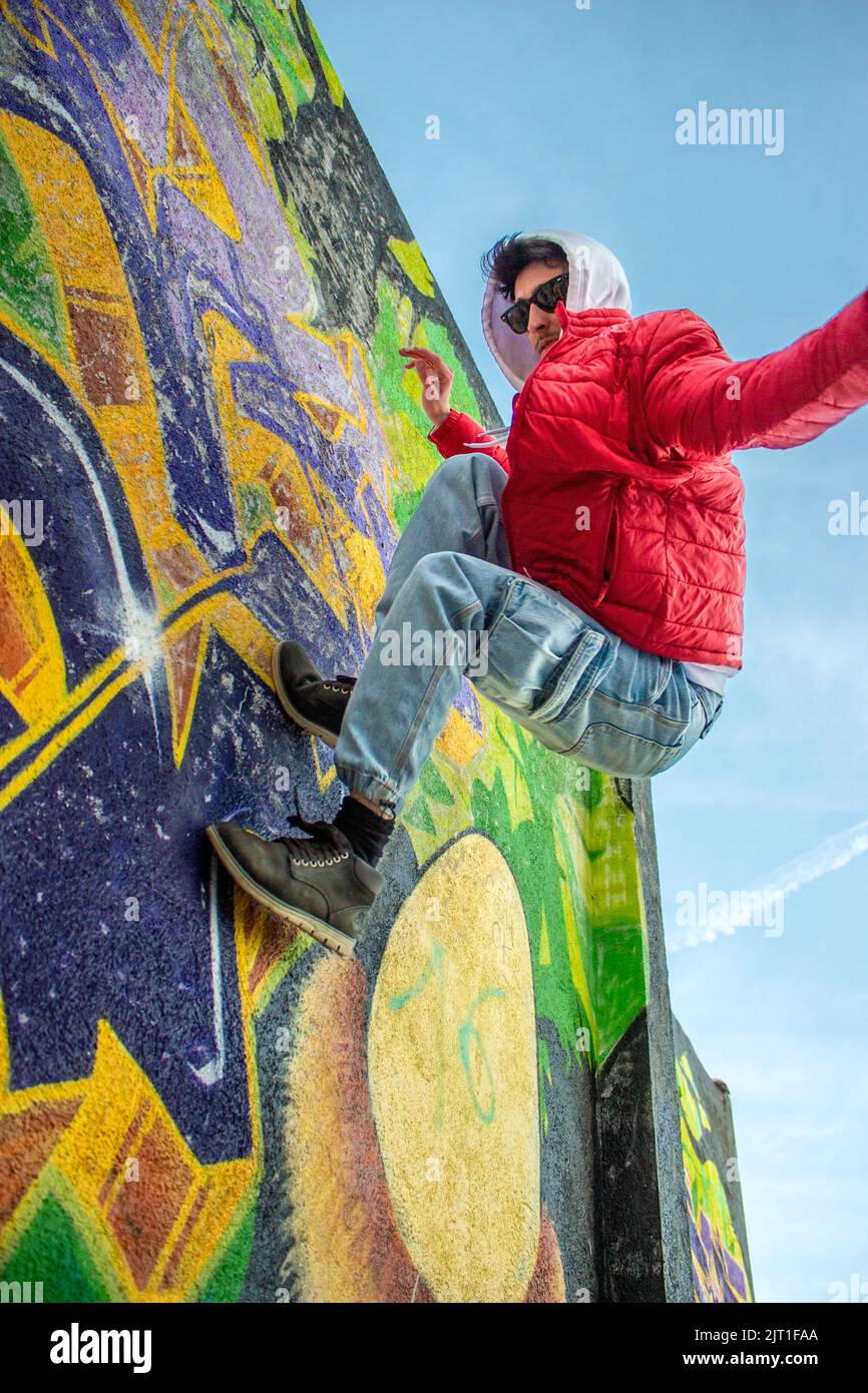 Elegante uomo caucasico che salta sui graffiti. Indossa occhiali da sole e una giacca rossa. Foto Stock