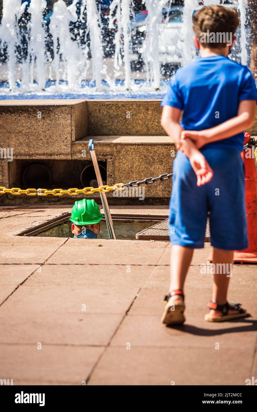Ragazzo bianco che guarda l'operaio di manutenzione delle fontane che esce dal sottosuolo a Savassi a Belo Horizonte, Brasile. Foto Stock