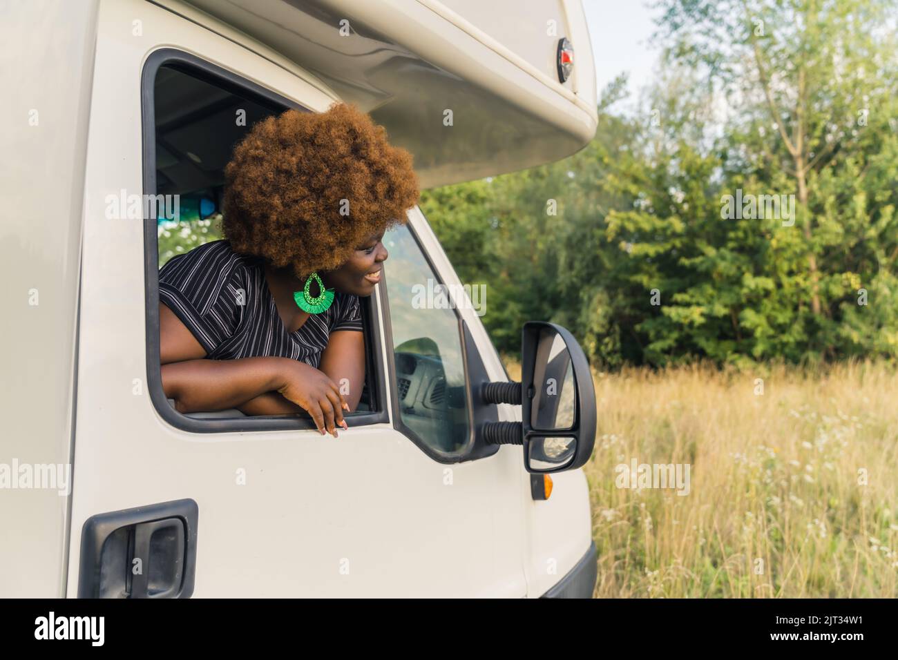 Giovane donna africana sorridente dalla pelle scura con capelli ricci che sporgono dalla finestra di un camper e che ammirano la natura pacifica intorno alla fine del suo viaggio. Foto di alta qualità Foto Stock