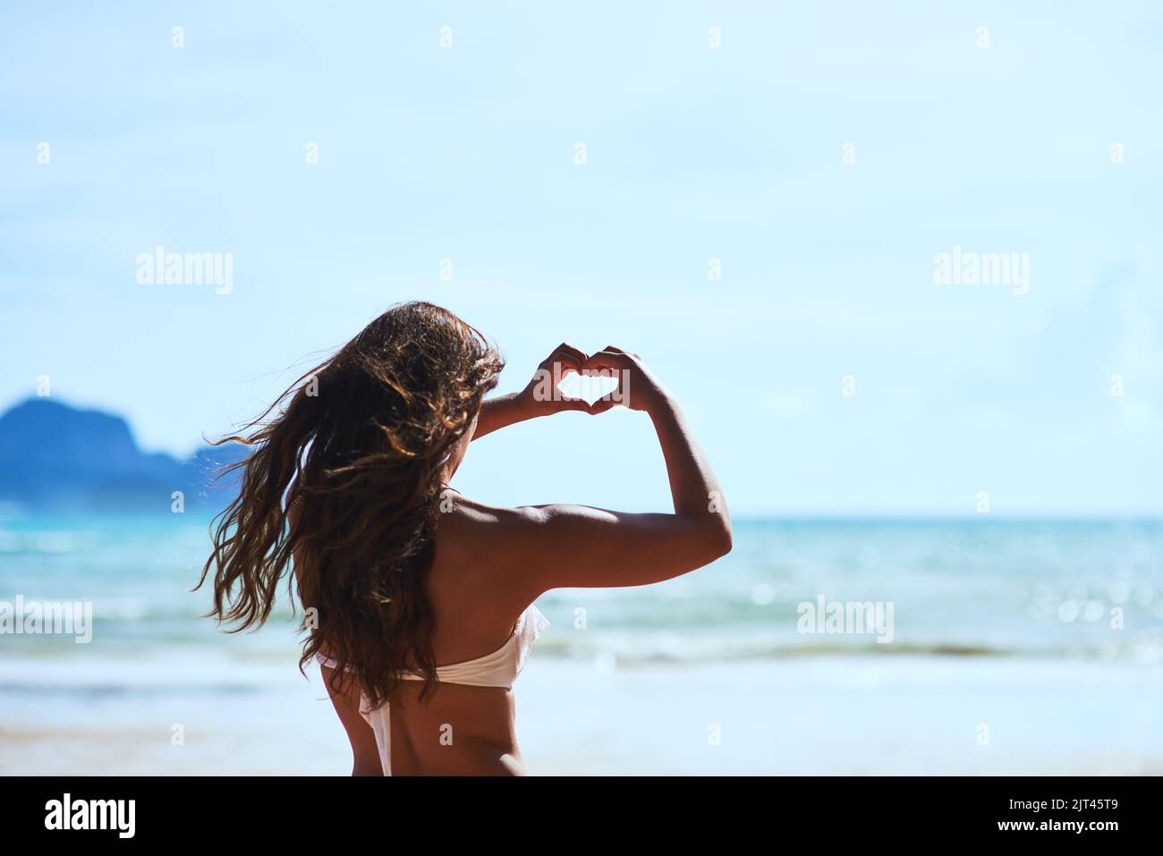 Che cosa non ama circa l'estate. Una giovane donna che forma un cuore mentre guarda il paesaggio alla spiaggia. Foto Stock
