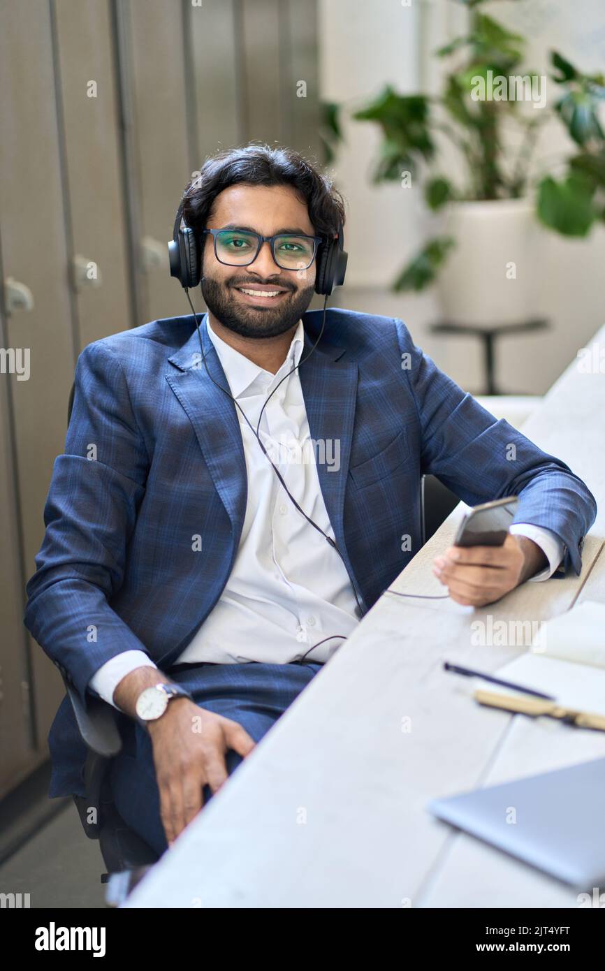 Uomo d'affari indiano che indossa le cuffie utilizzando il telefono al lavoro. Verticale Foto Stock